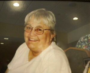 Smiling elderly woman with glasses, short gray hair, in a light pink shirt. Lit room setting.