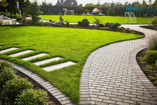 Well-manicured lawn with stone path winding through, framed by bushes, with a soccer field in the background. Well-manicured lawn with stone path winding through, framed by bushes, with a soccer field in the background.