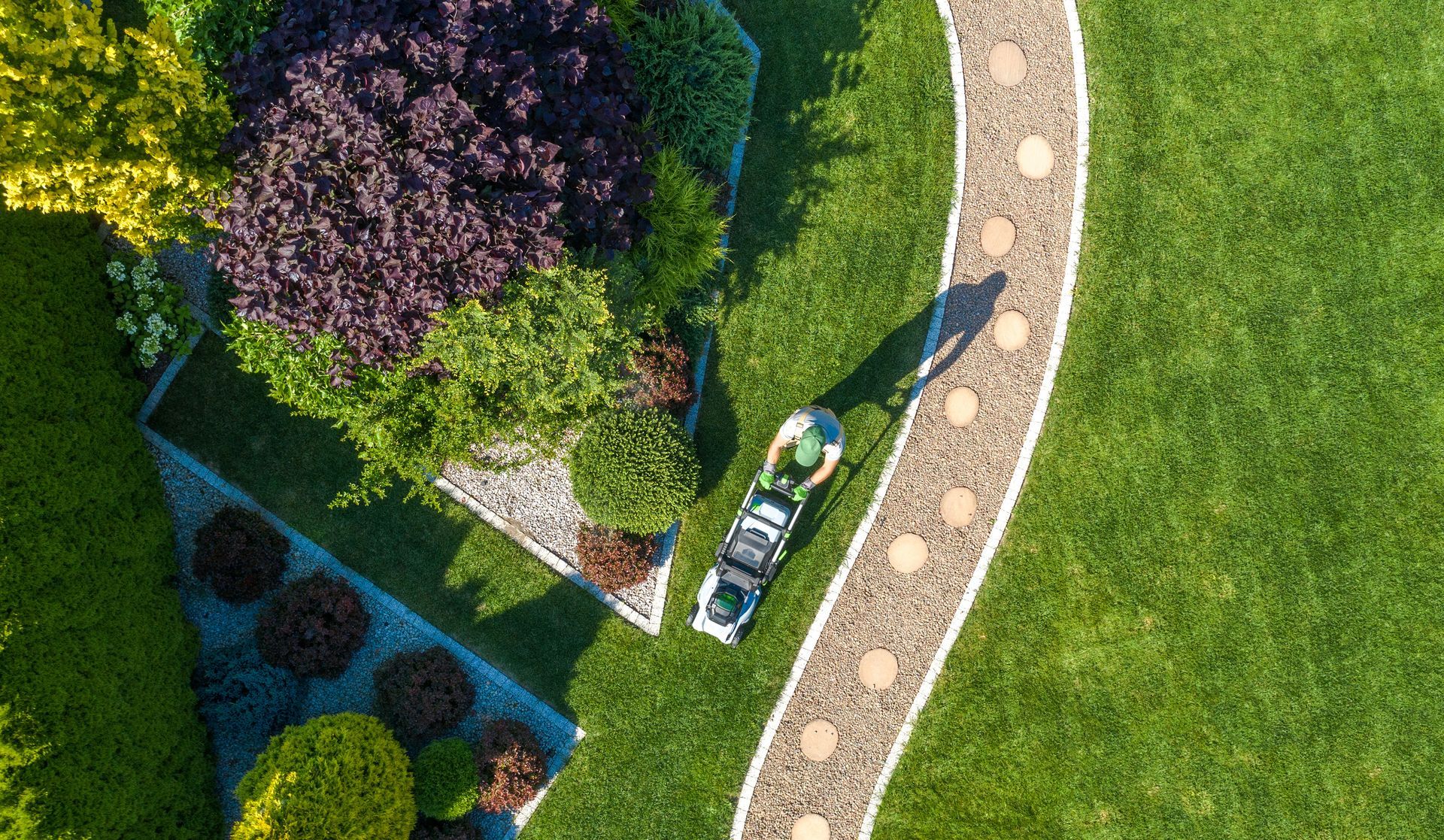 Overhead view of a garden with a winding stone path, green lawn, flower beds, and a lawnmower. Overhead view of a garden with a winding stone path, green lawn, flower beds, and a lawnmower.