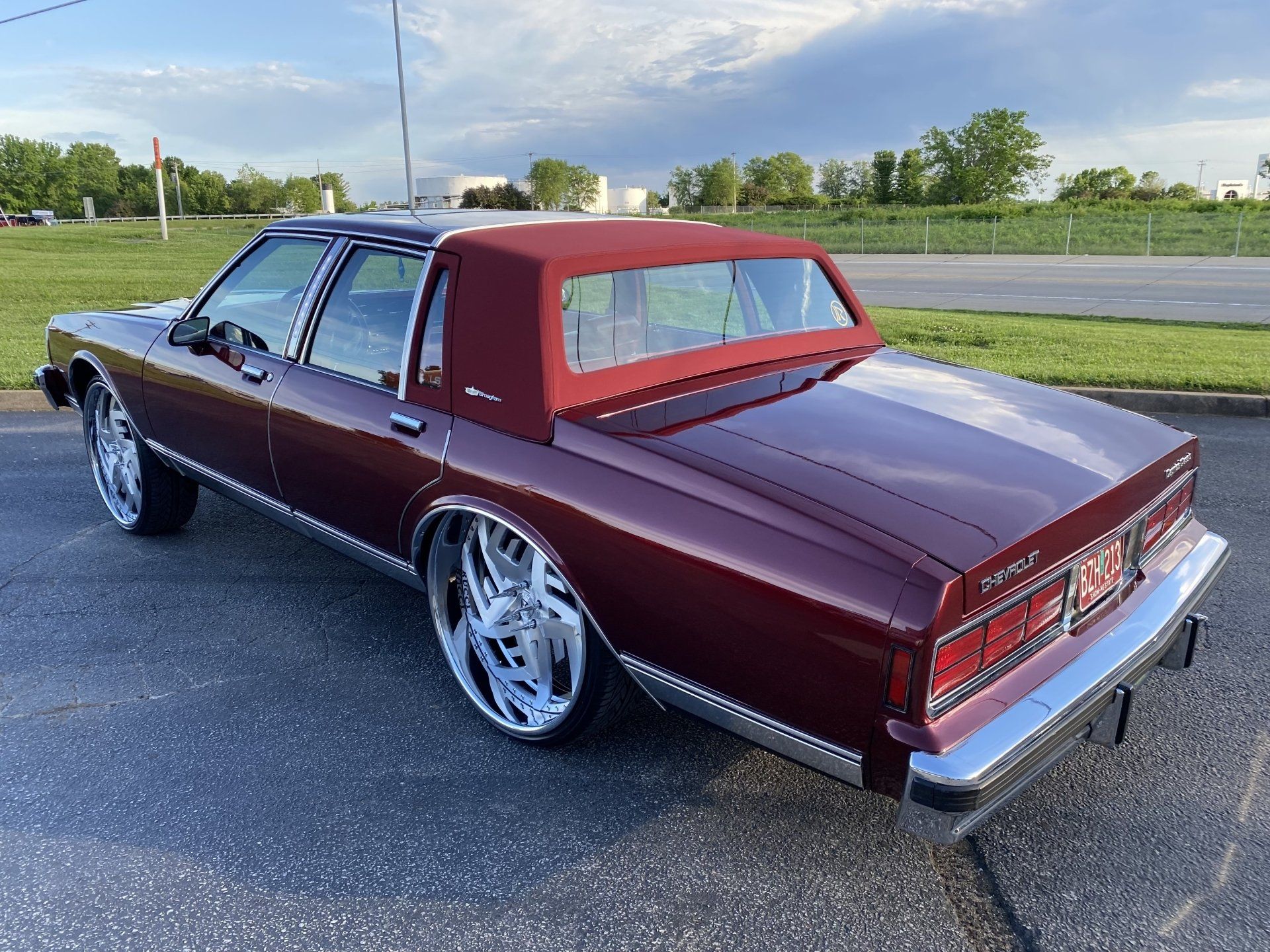 Maroon classic coupe with white vinyl roof parked on an asphalt lot by a grassy field