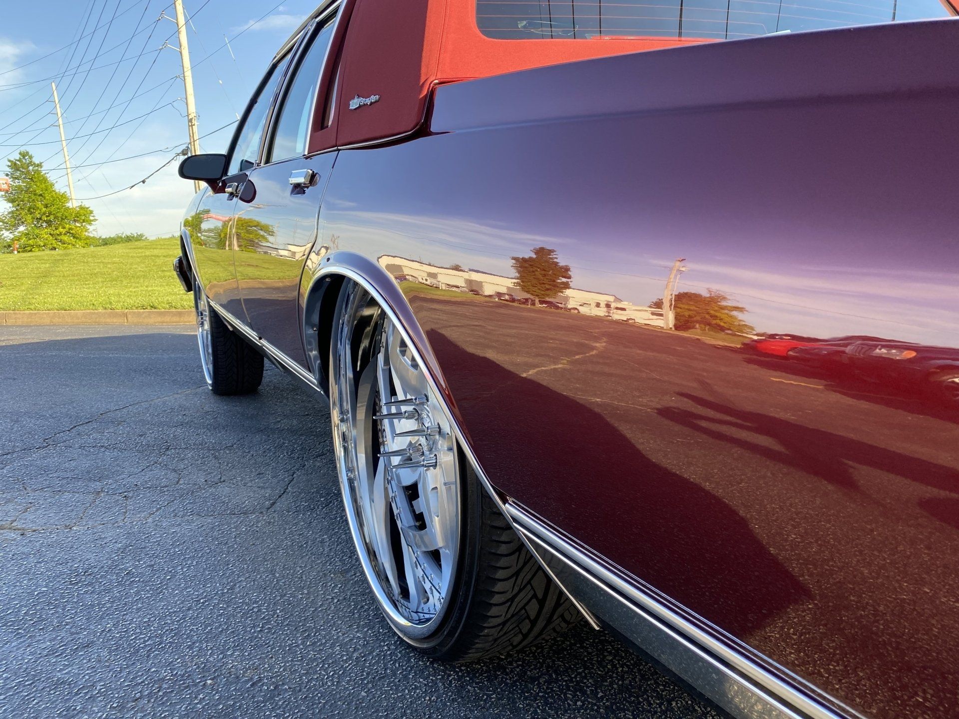 Low-angle view of a maroon classic car parked on pavement, with polished chrome wheels reflecting sunlight.