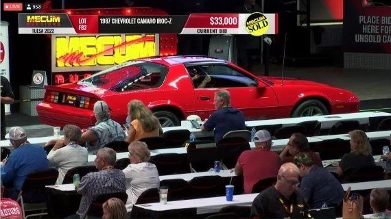 Red classic sports car on stage at auction, with seated audience in foreground and bid price displayed above