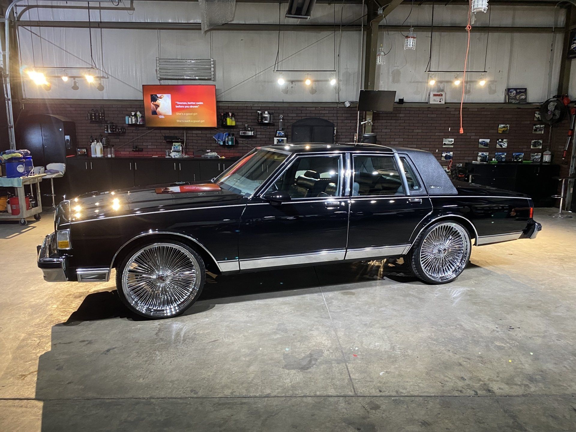 Black classic sedan with chrome rims parked in a dim garage or workshop under bright lights