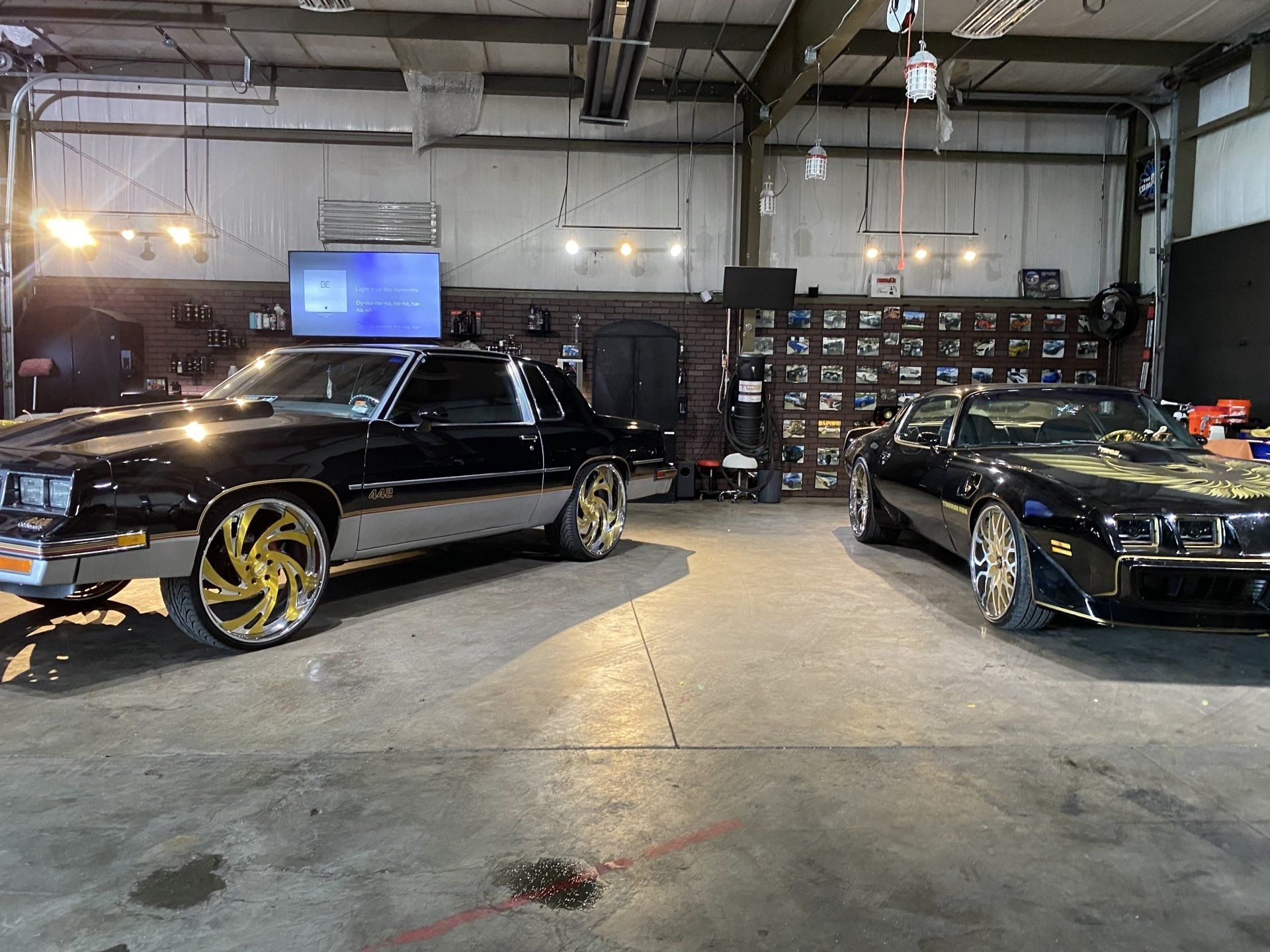 Two black custom cars parked inside a garage with bright overhead lights