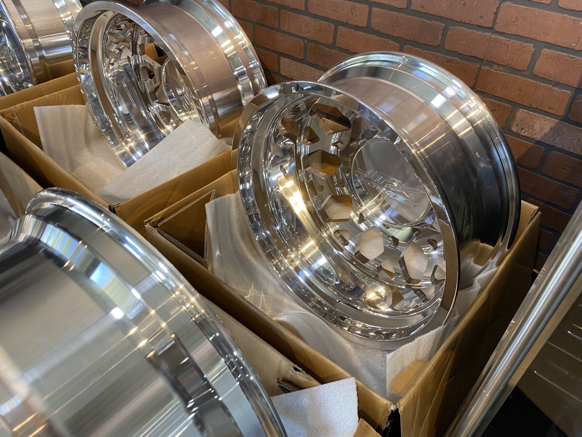 Polished chrome car rims stacked in cardboard boxes against a brick wall.