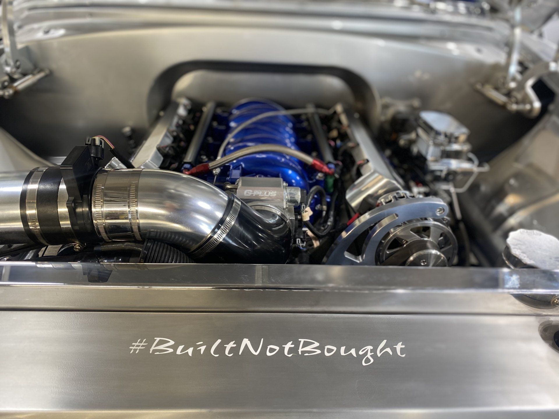 Modified car engine bay with polished intake piping and blue hoses, viewed from the front trunk area