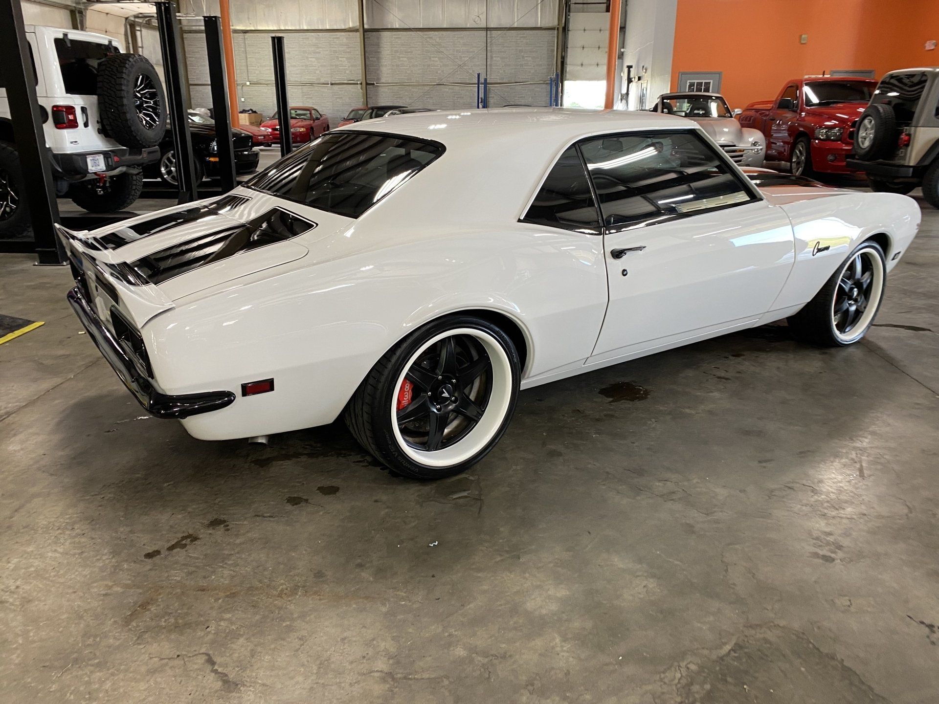White classic muscle car in a garage, shown from the rear side with black wheels and hood vents.