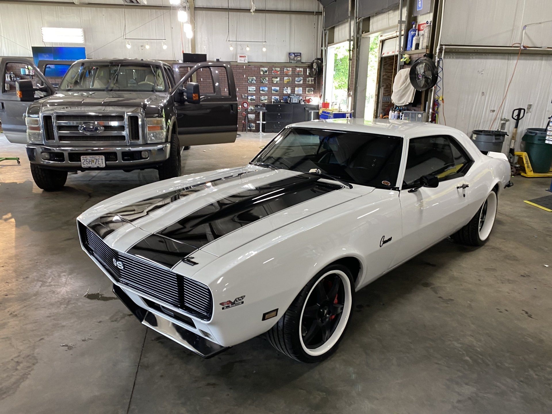 Classic white muscle car with black stripes parked inside a garage next to a pickup truck