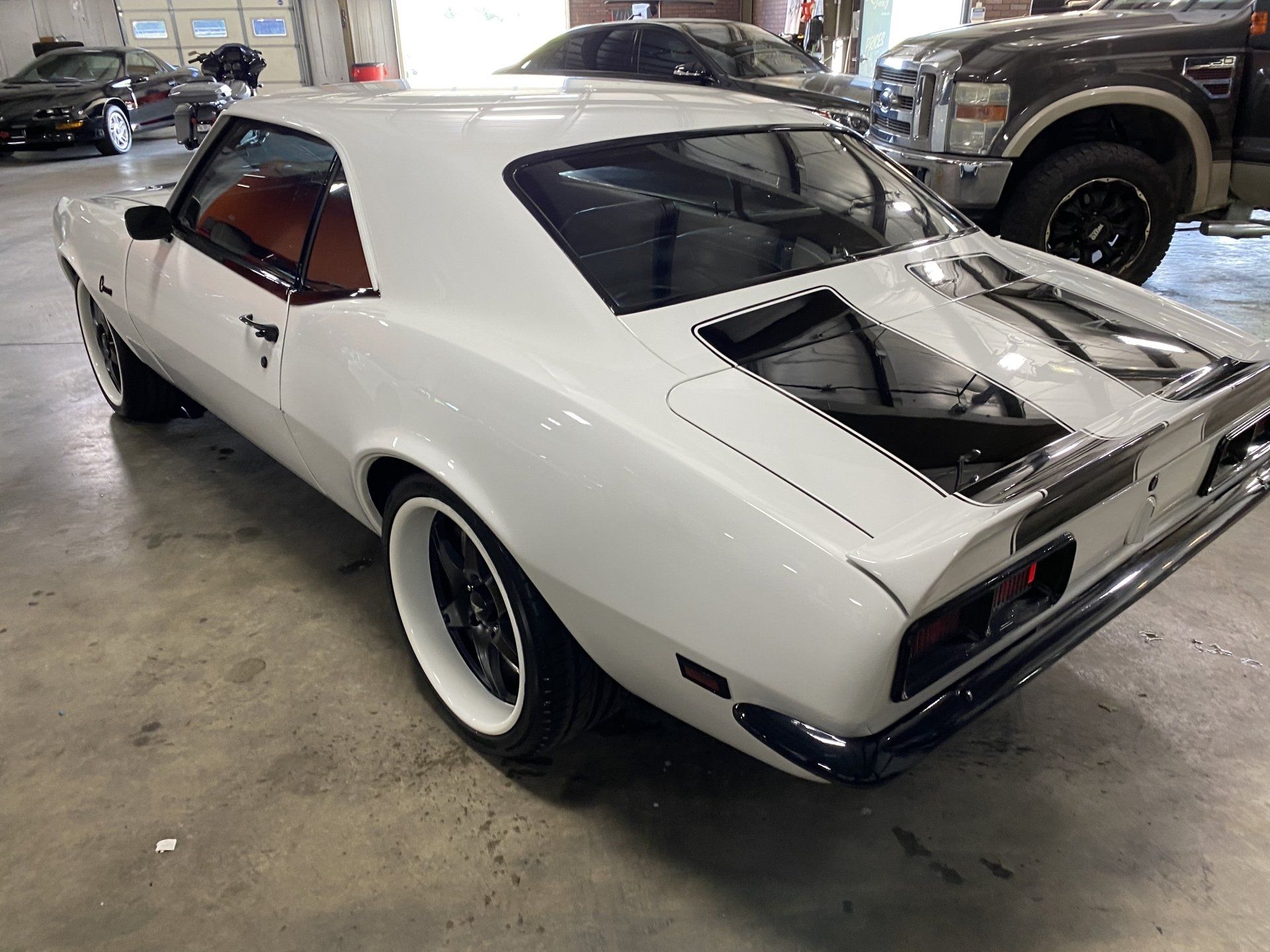 White classic muscle car in a garage, viewed from the rear with exposed engine bay and black racing stripes.