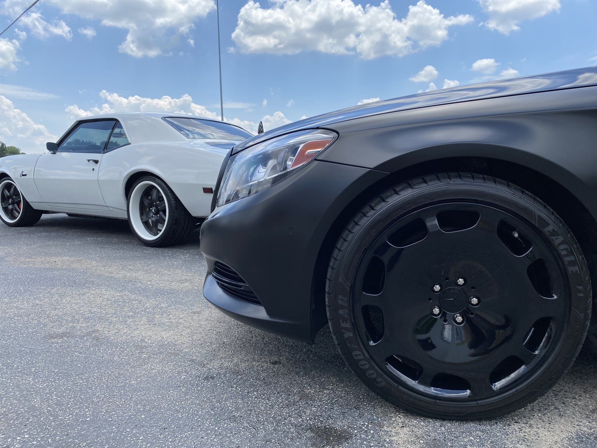 Two custom cars parked on gravel under a cloudy blue sky, one white and one black in the foreground