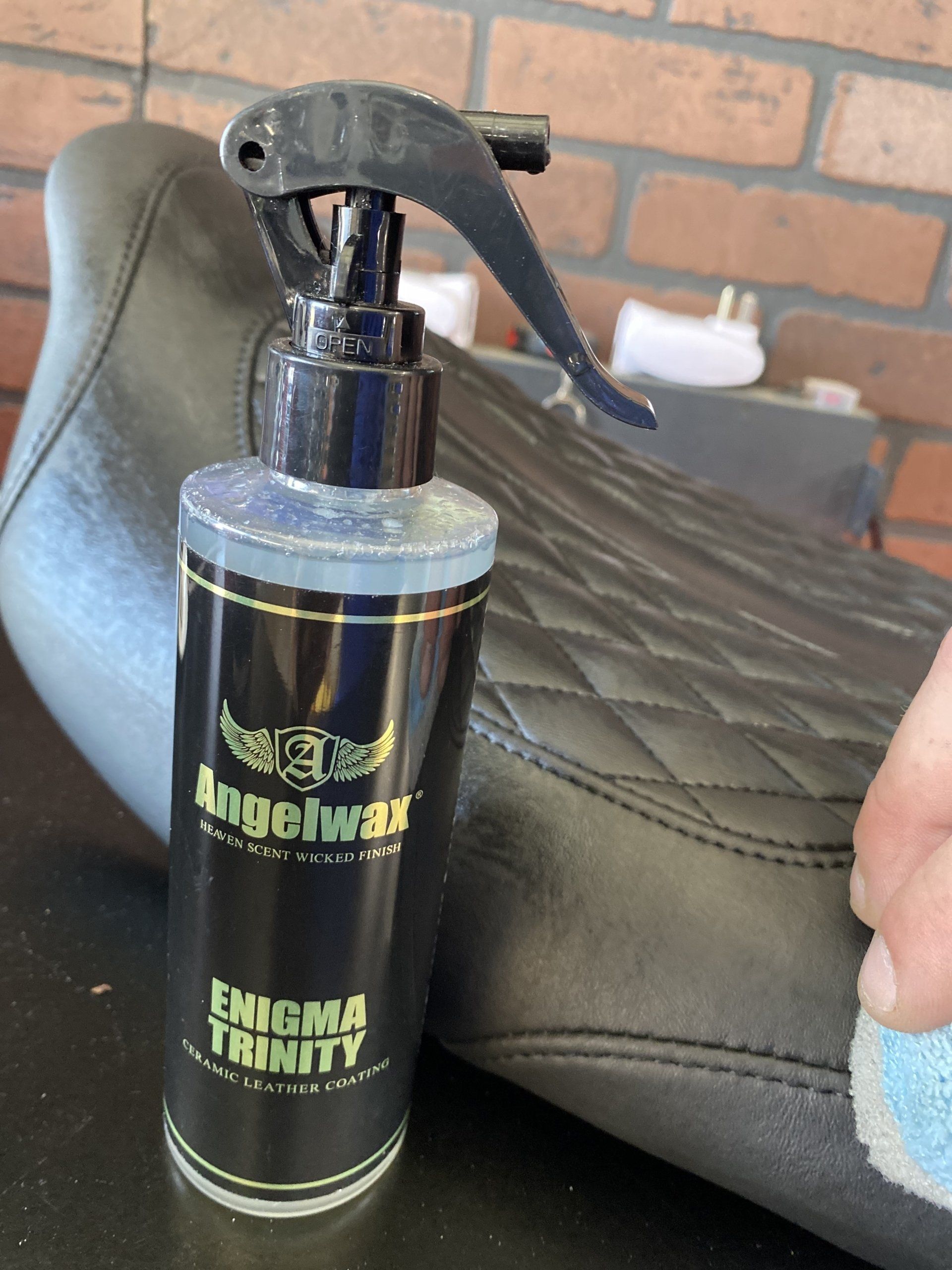 Black spray bottle with green label on quilted bench, held by a hand near a brick wall.
