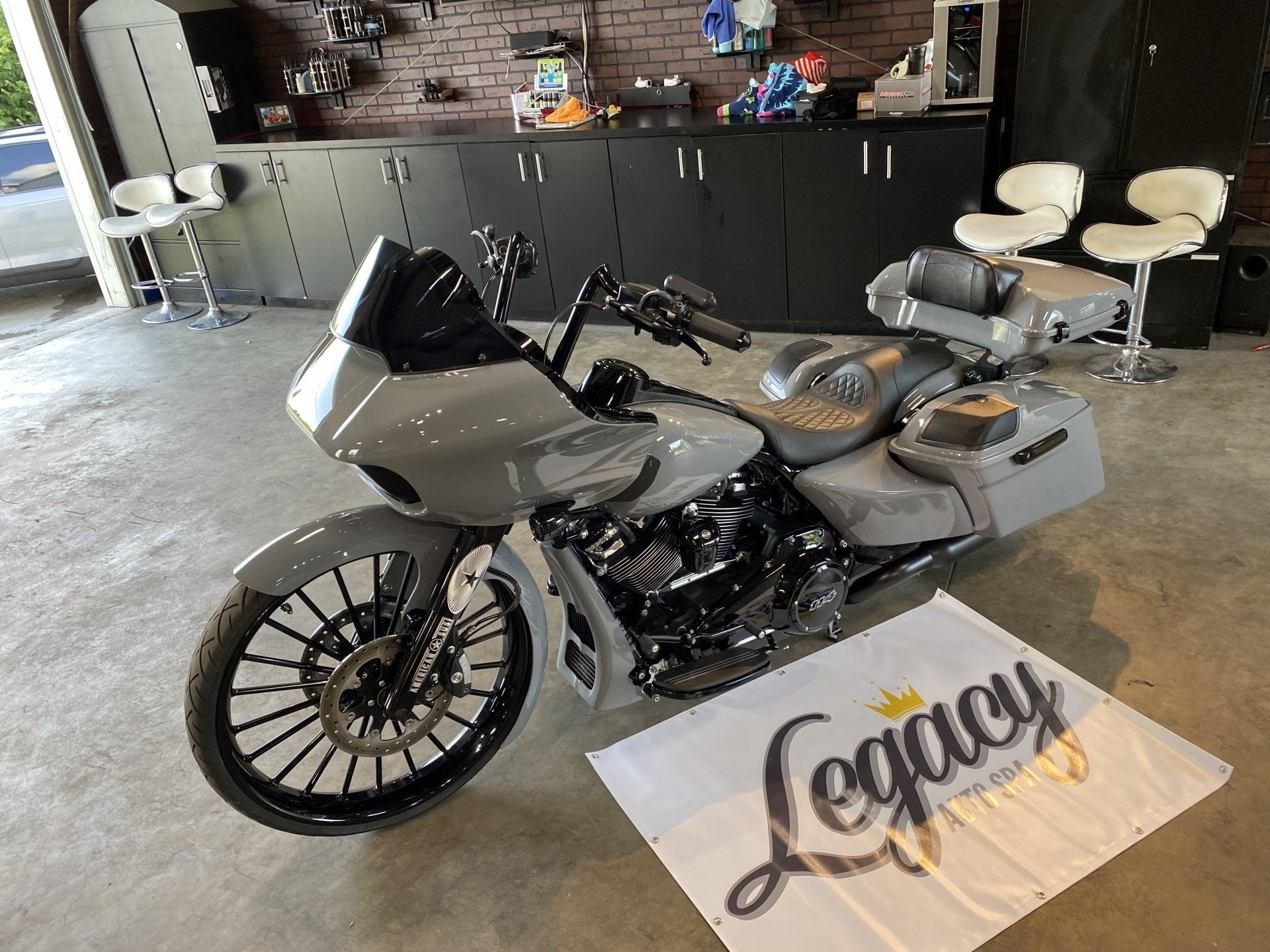 Silver custom motorcycle on display in a garage, with a black cabinet wall and stools behind it.