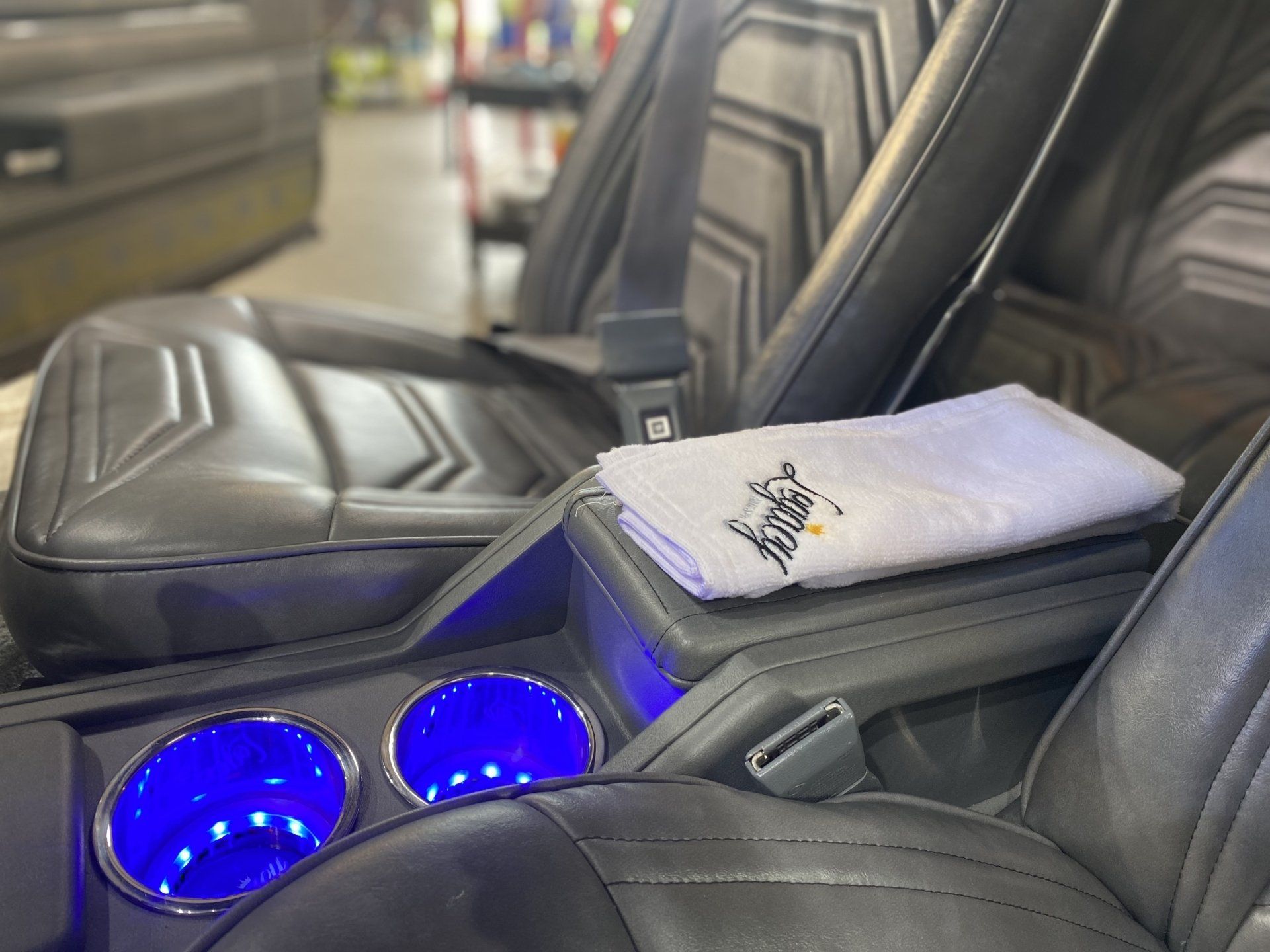 Car armrest with glowing blue cup holders and a folded white towel on black leather seats