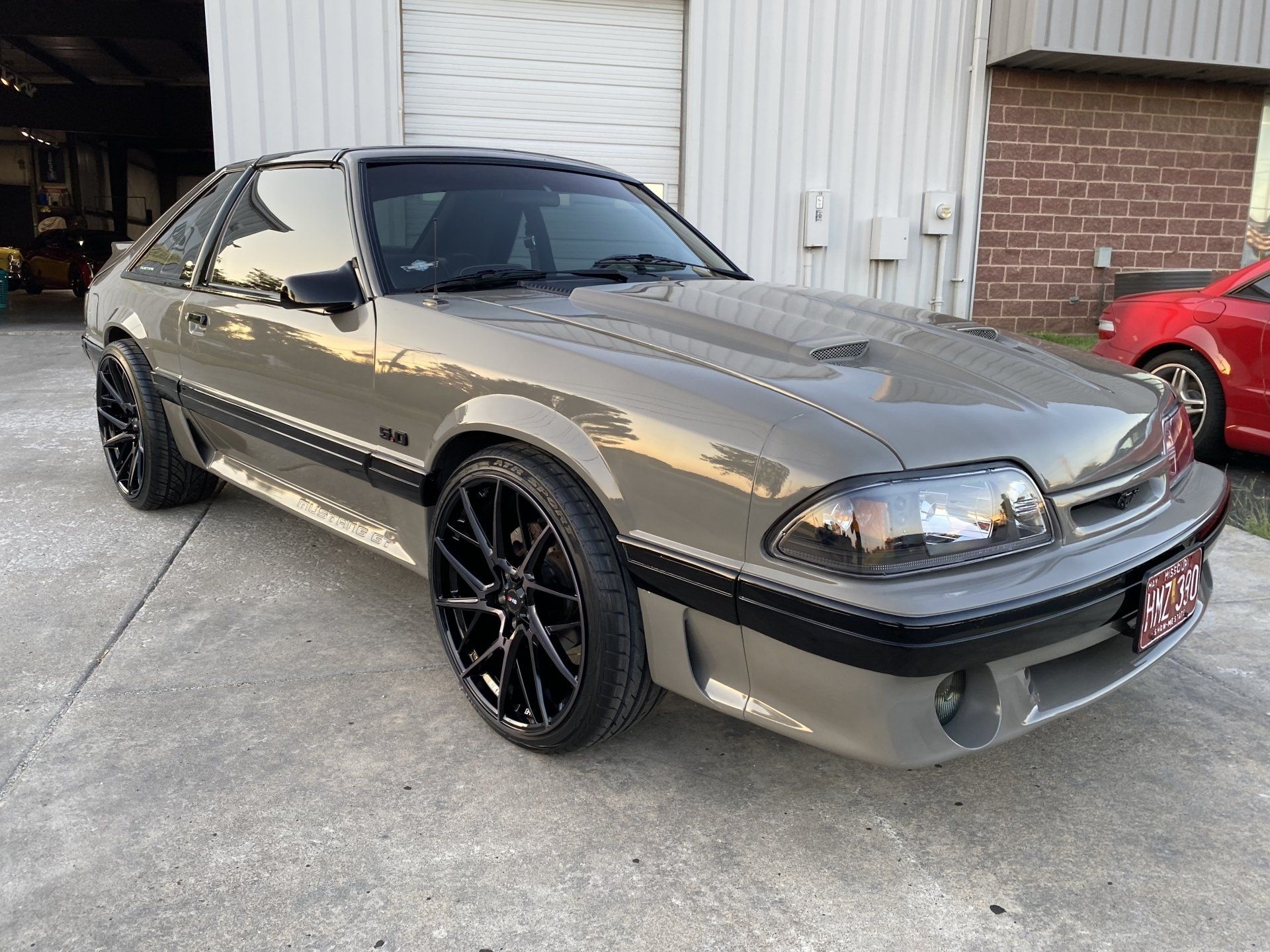 Gray sports coupe parked outside a garage, with black rims and a red car in the background