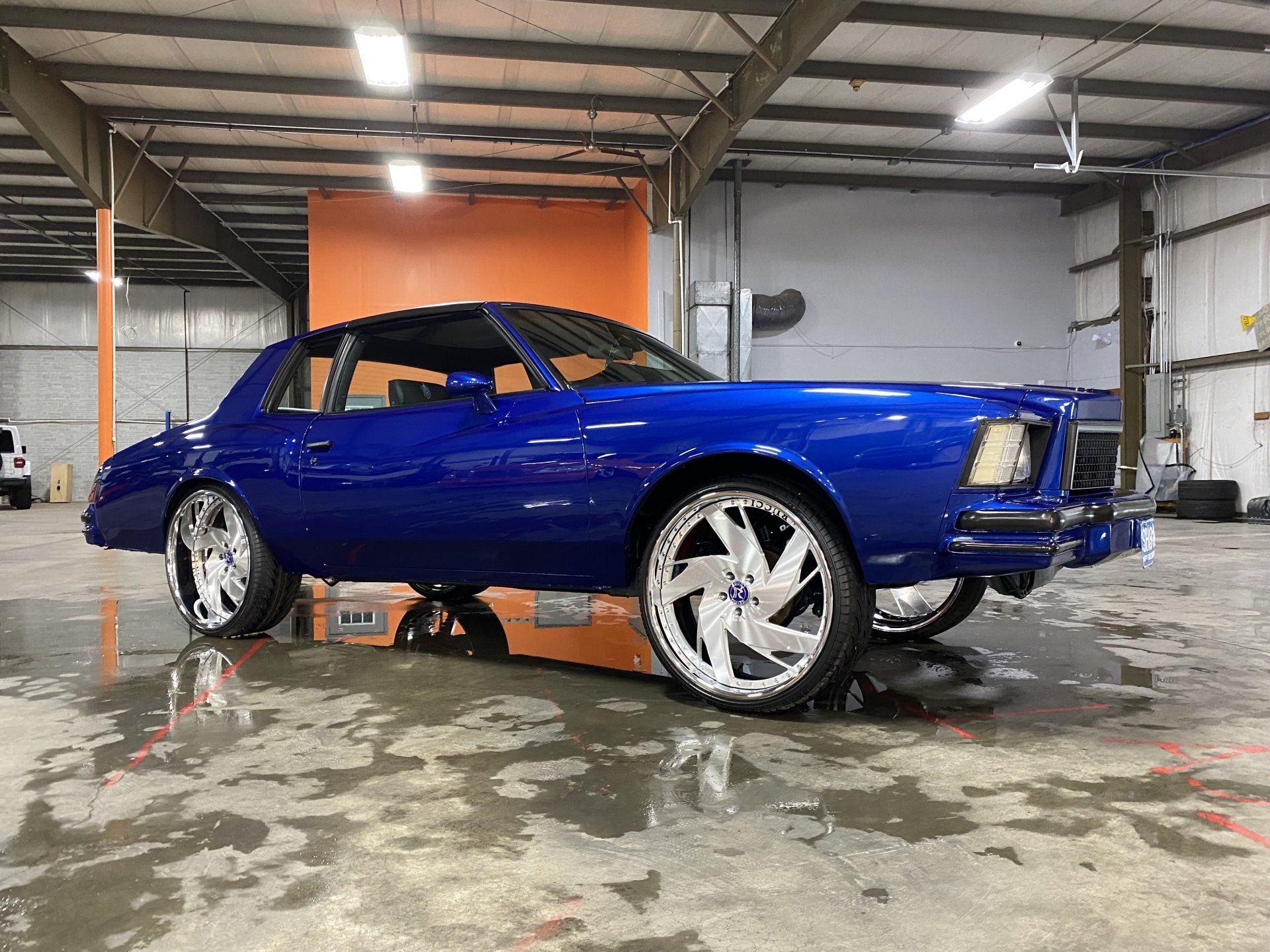 Blue classic car in a warehouse, elevated on a lift with chrome wheels and wet floor beneath.