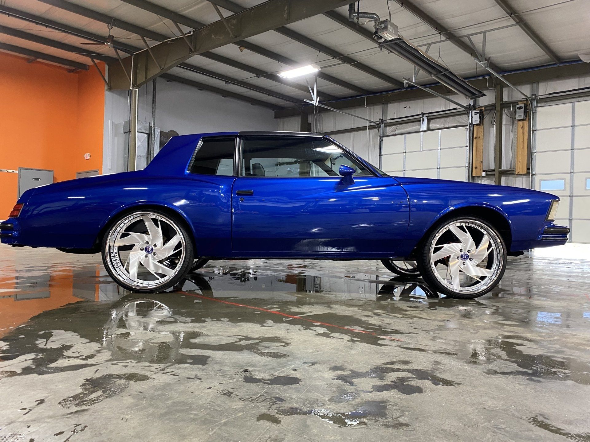 Blue classic coupe parked in an industrial garage, with chrome wheels and a wet reflective floor.
