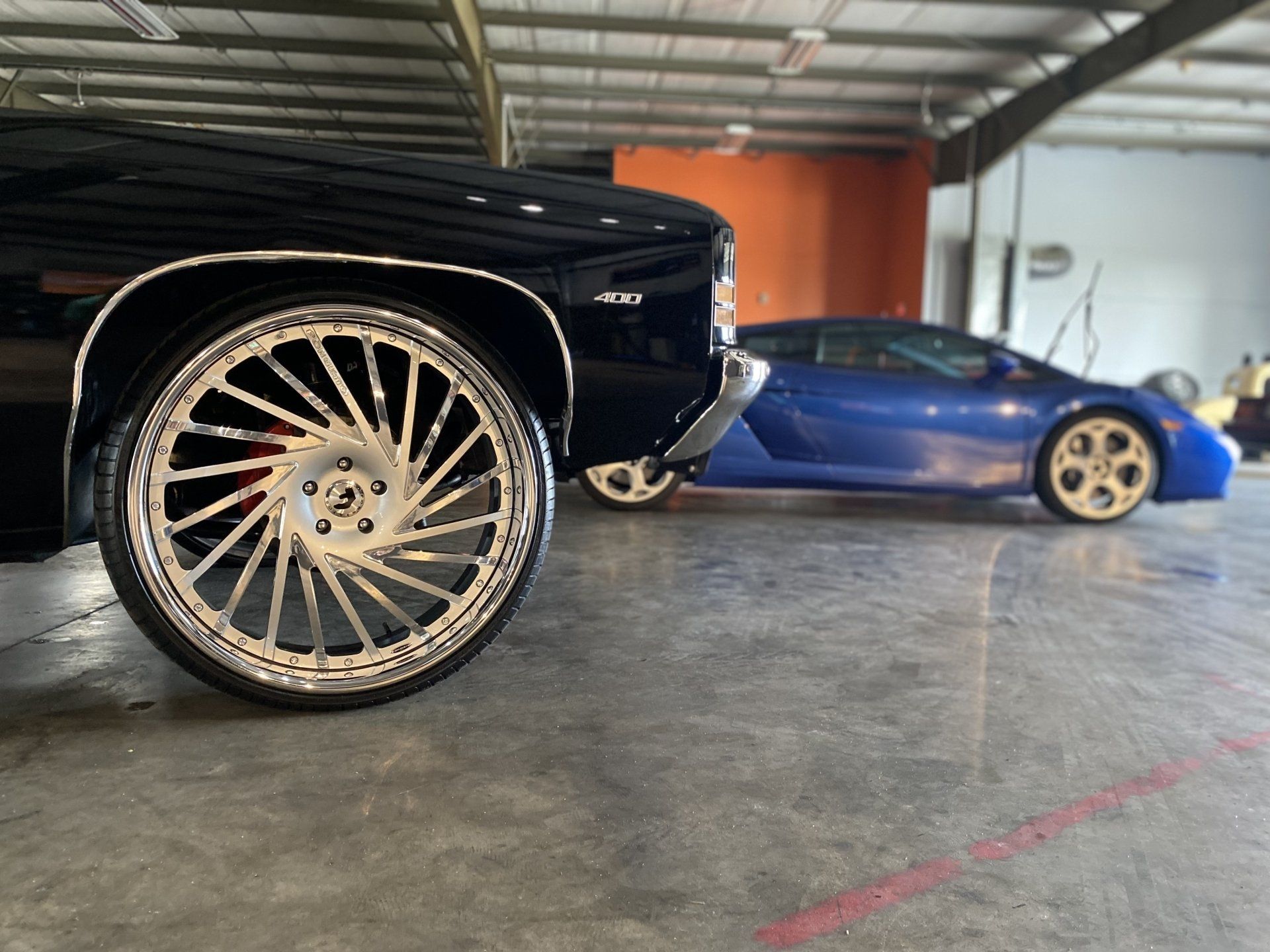 Black custom car with large chrome wheel in foreground, blue sports car behind in a garage