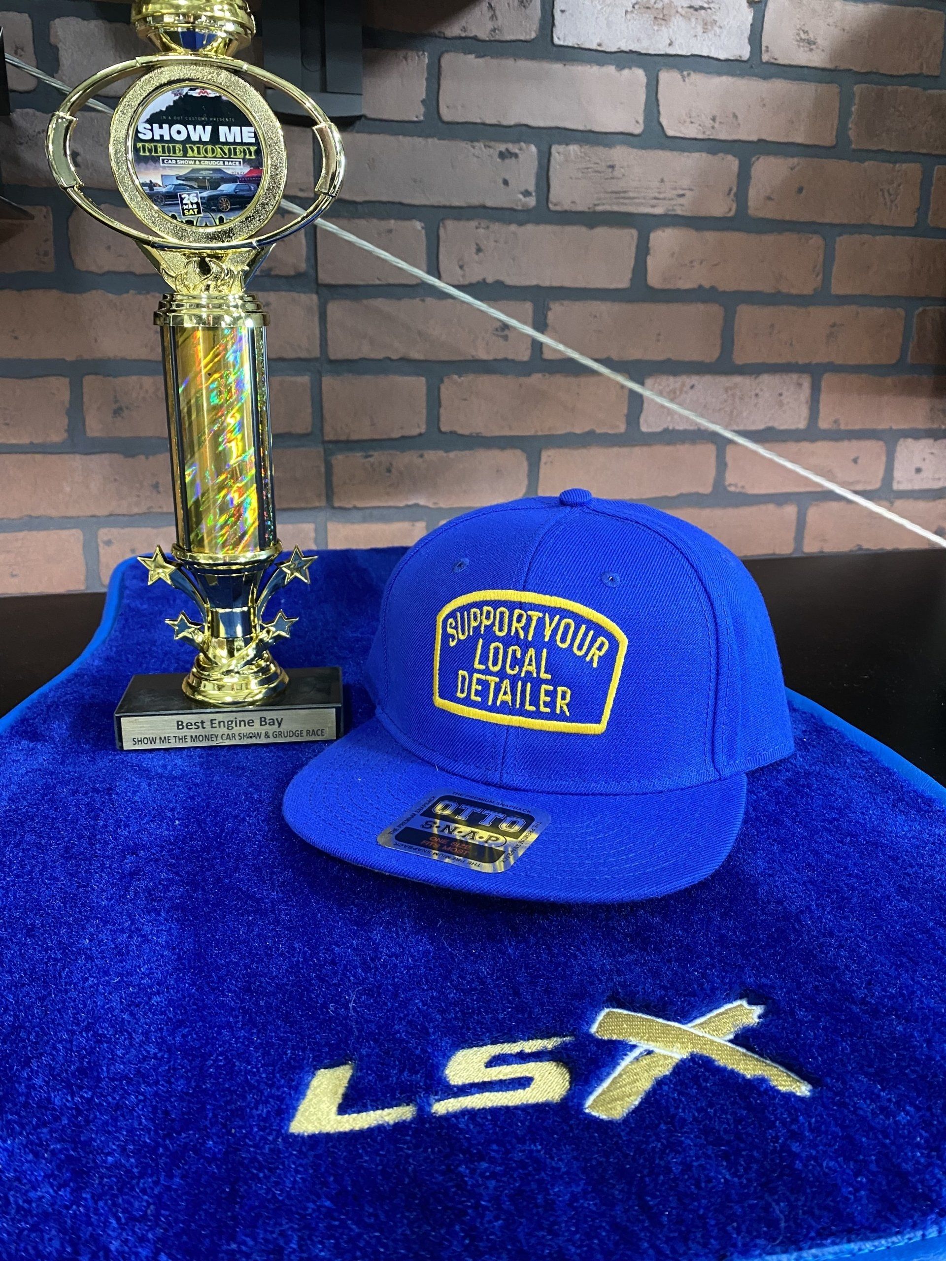 Blue LSX cap and towel beside a gold trophy on a brick backdrop