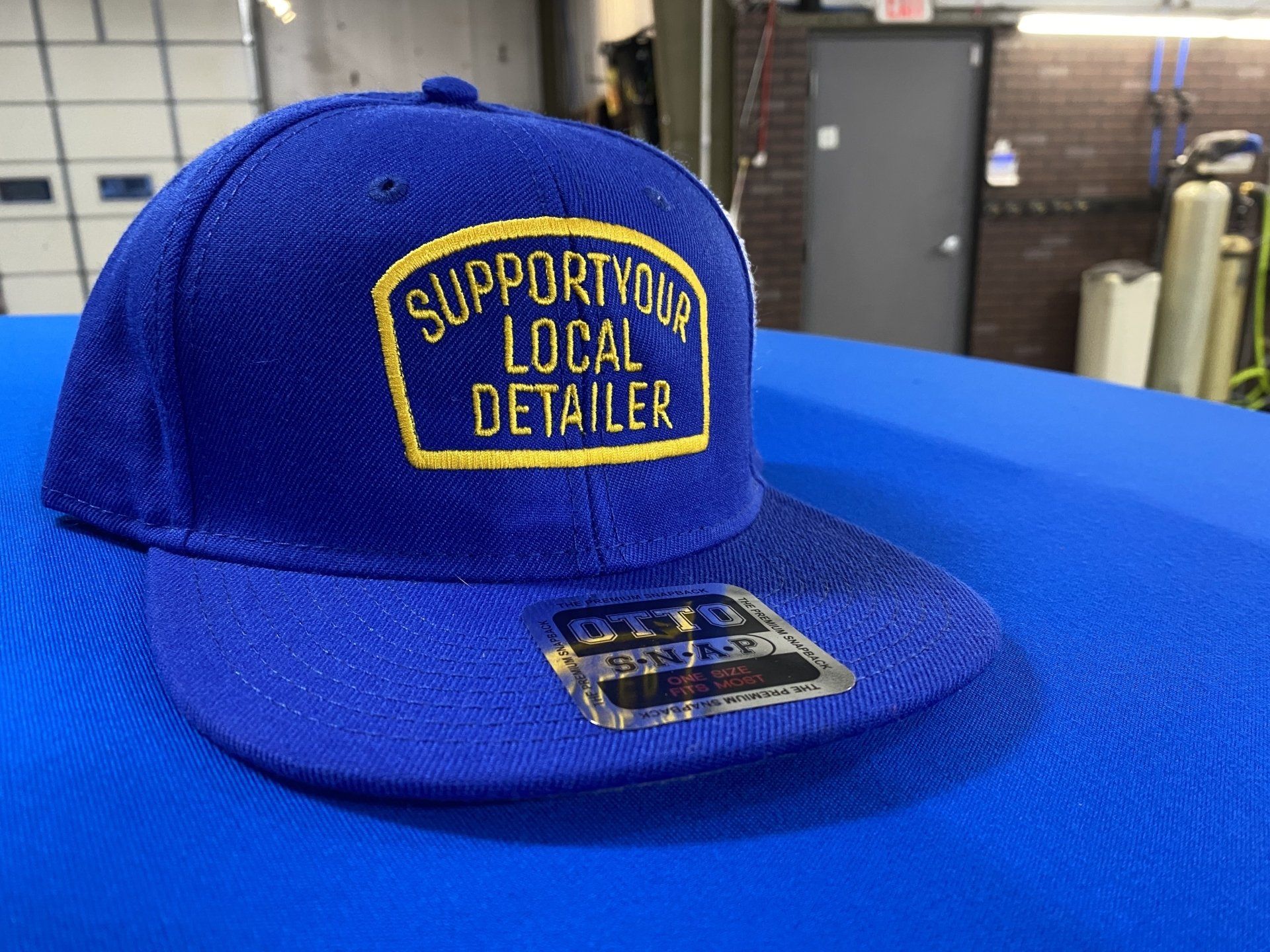 Blue cap with “Support Your Local Detailer” patch on a blue surface in a workshop setting