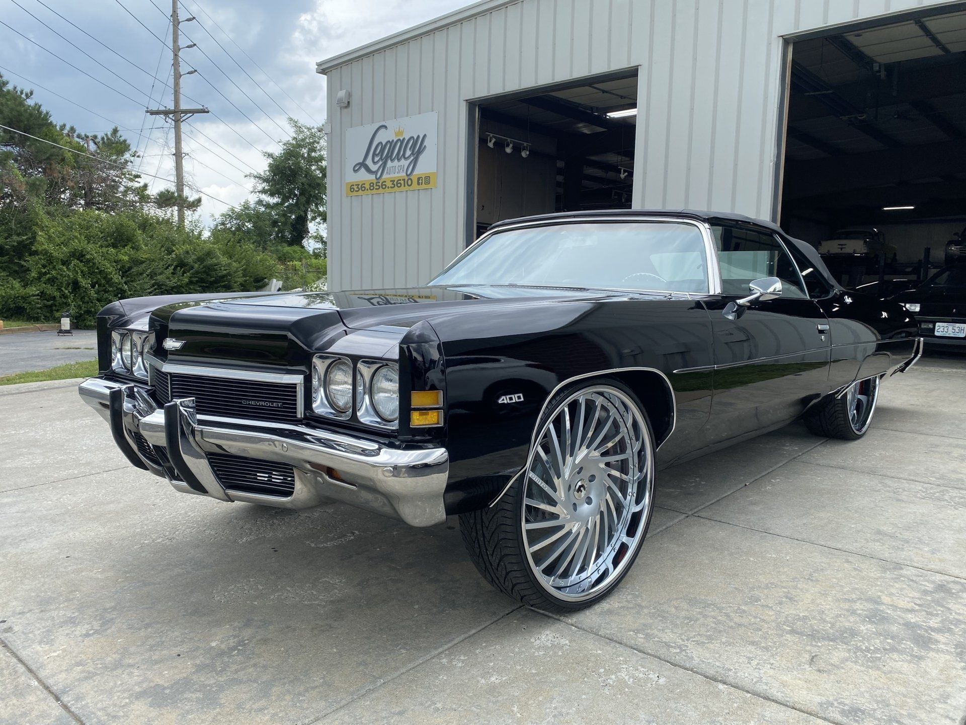 Black classic convertible with oversized chrome rims parked outside a garage.