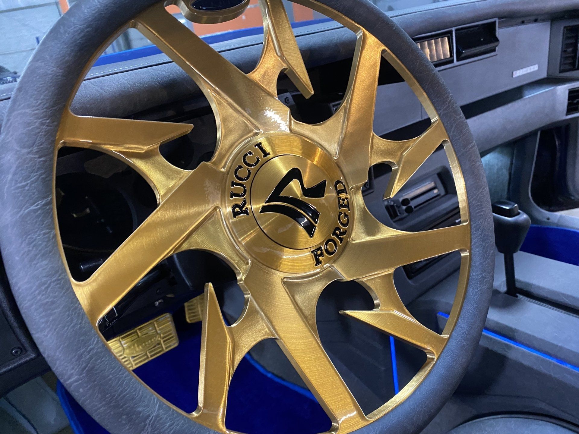 Gold custom wheel rim with black tire, showing a stylized logo inside a car interior.