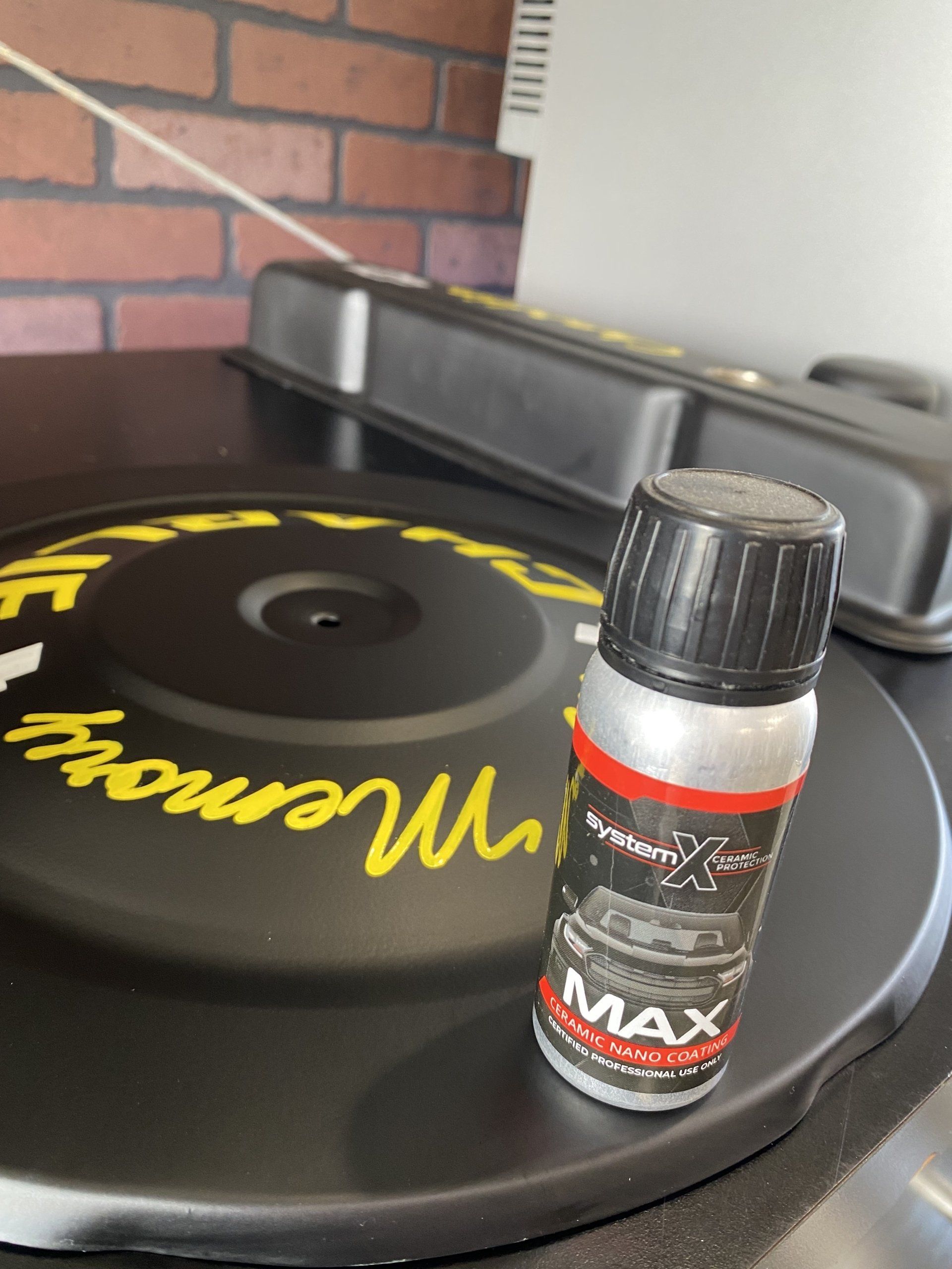 Small bottle of MAX motorcycle cleaner on a black turntable with yellow lettering, against a brick wall.