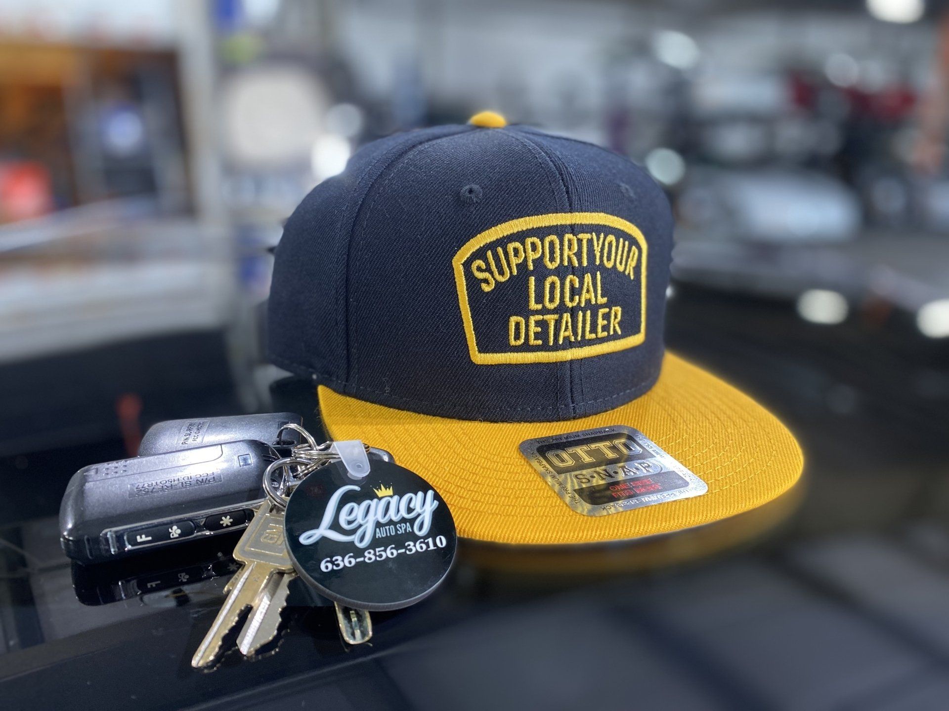 Navy and gold “Supporting Local Detailer” cap beside car keys and a black key fob on a table