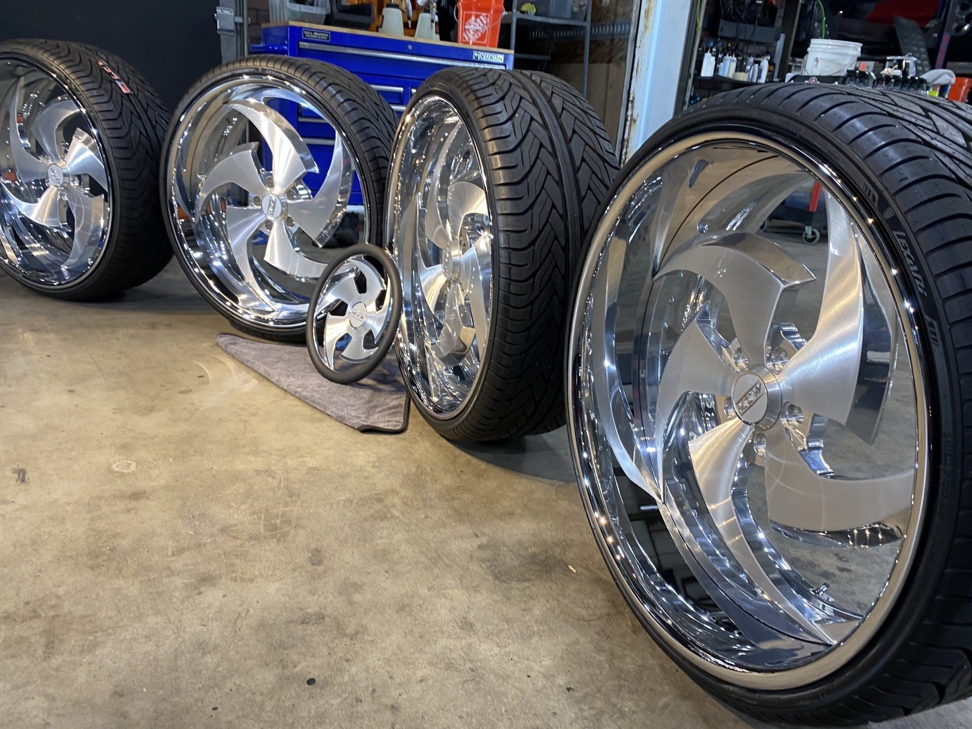 Custom chrome rims and tires lined up on a garage floor, reflecting the workshop lights.