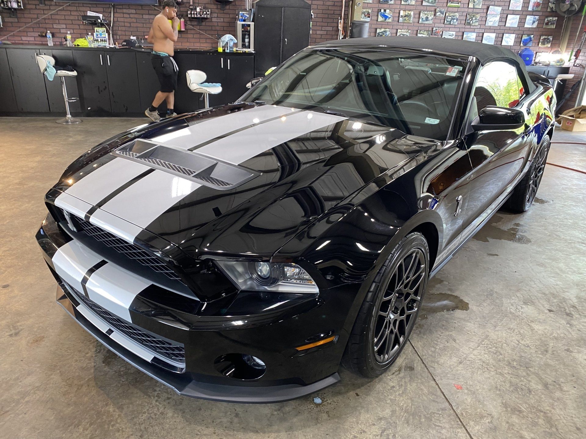Black sports car with white racing stripes in a garage, with people working in the background