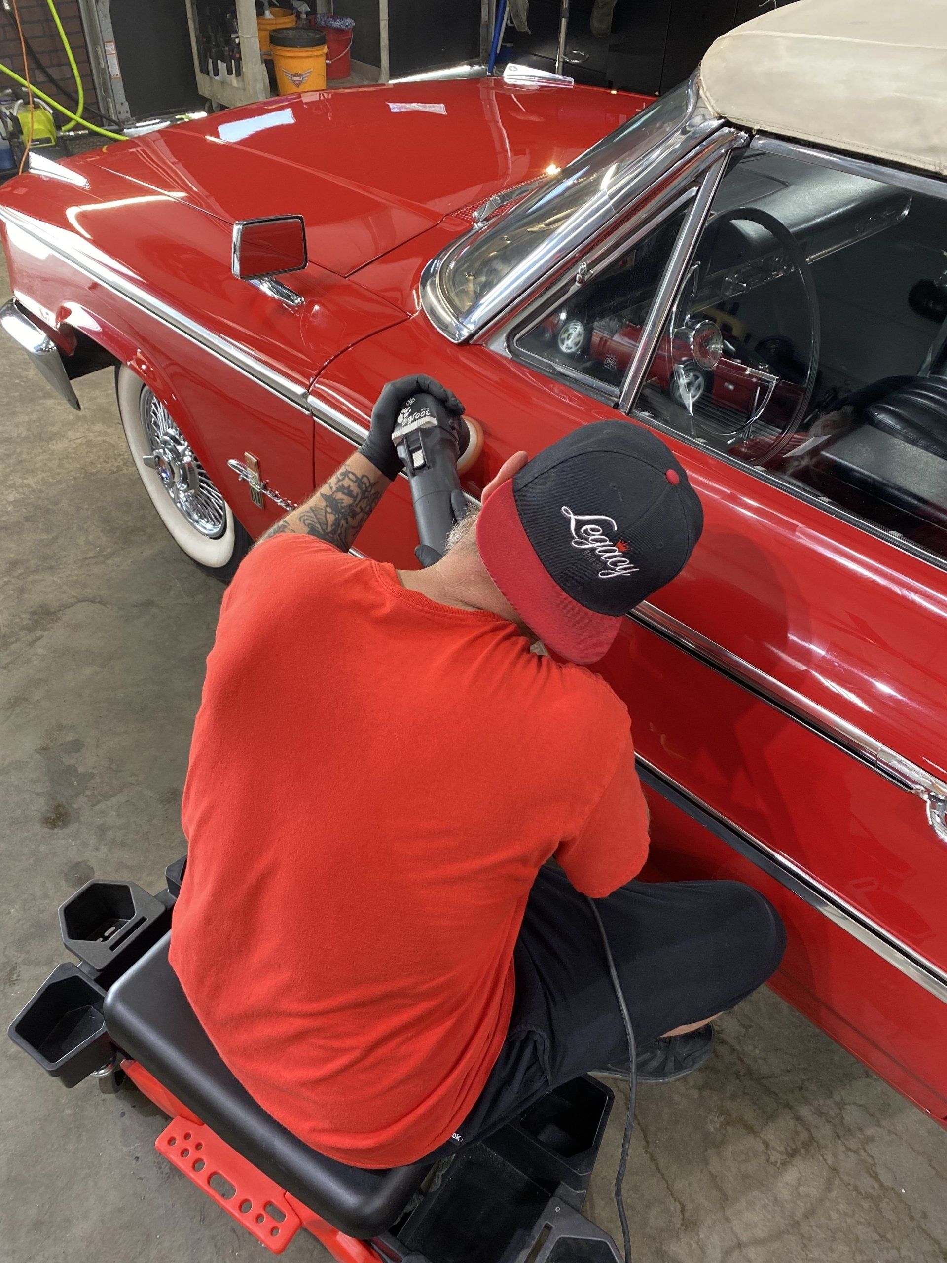 Person polishing a red classic car door in a garage, wearing a black cap and red shirt.