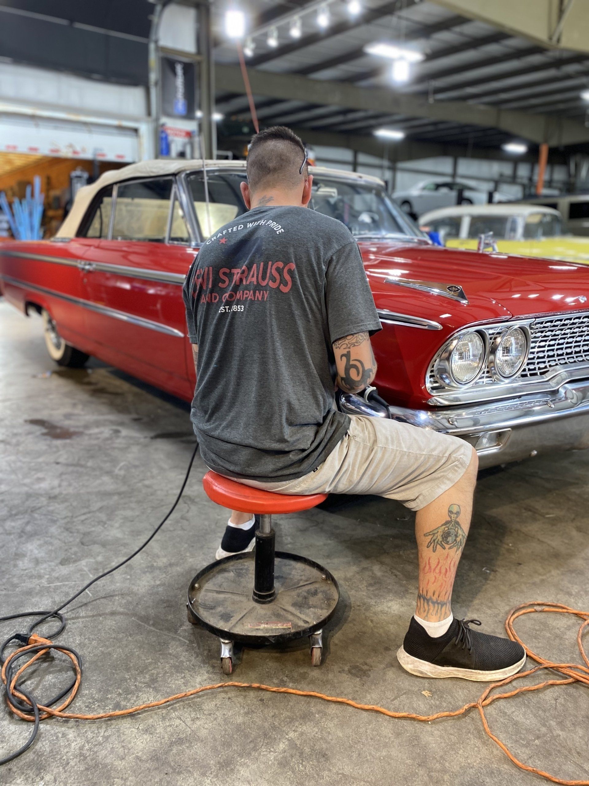 Man polishing a red classic car in an auto shop, seated on a rolling stool.