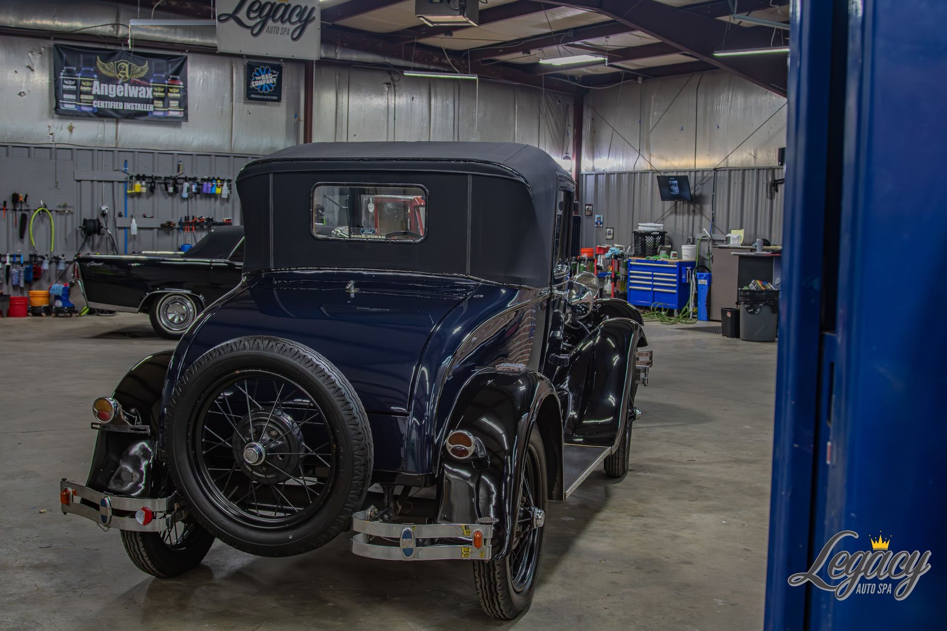 Vintage dark blue car in a workshop garage, viewed from the rear with tools and lifts in the background