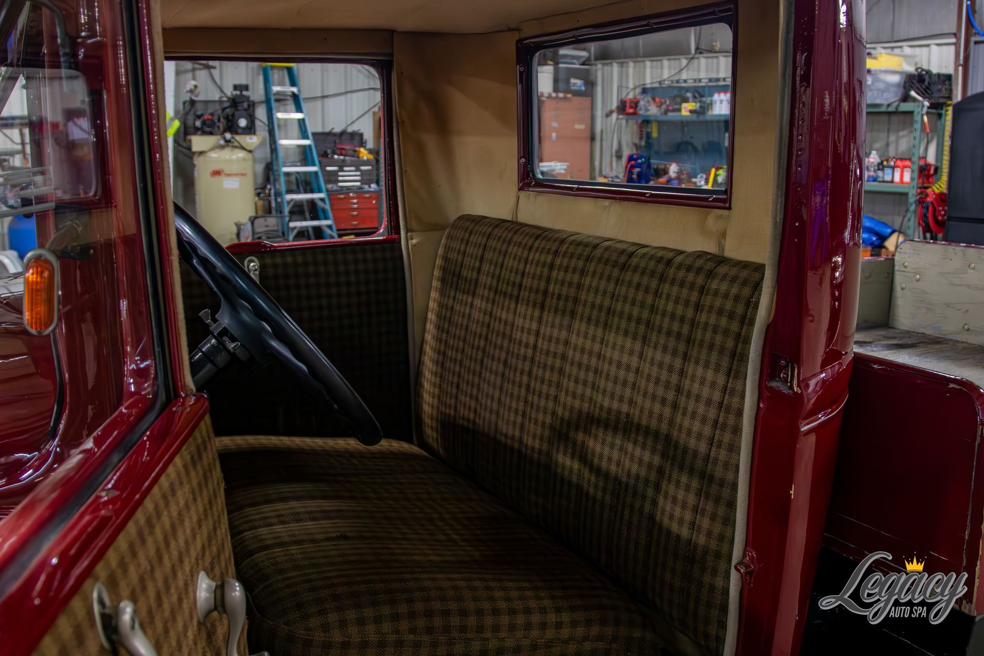 Inside a vintage car interior with plaid bench seats and windows into a garage workspace.