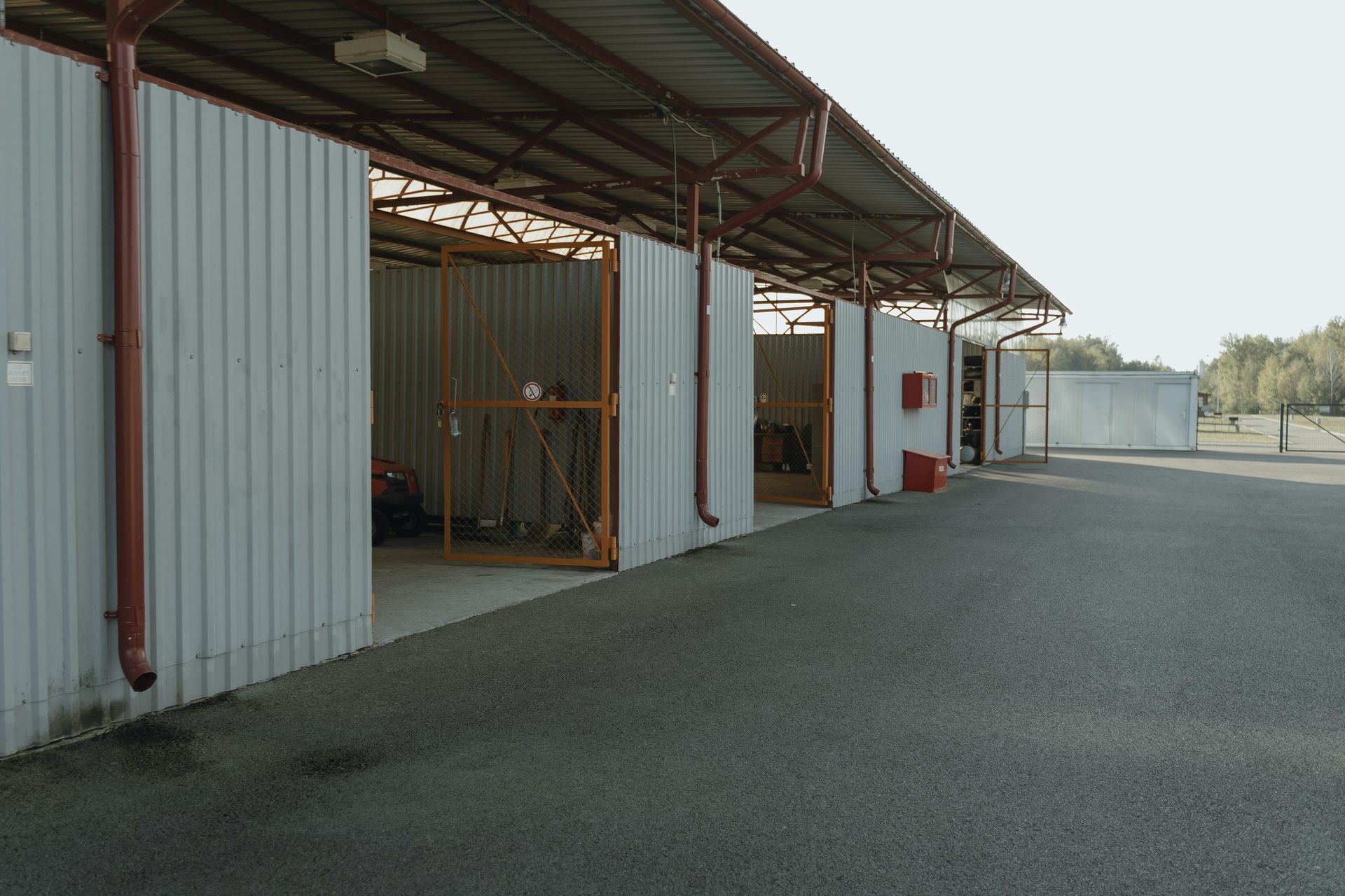 Metal storage sheds under a covered structure, asphalt surface, clear sky.