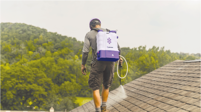 Worker on a rooftop using a backpack sprayer, with green trees in the background.