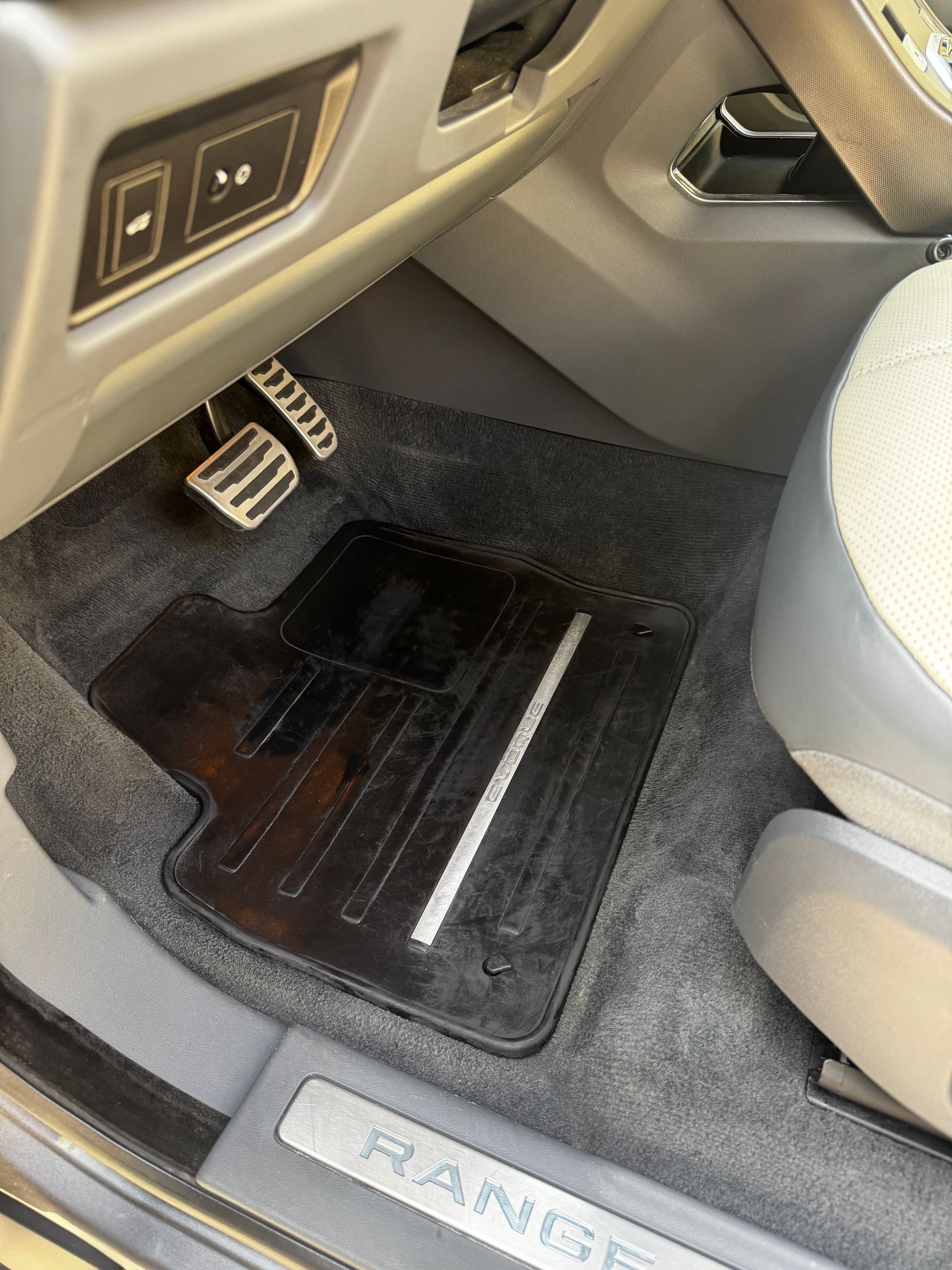 Car floor mat and pedals in a tan vehicle interior, with a Range Rover door sill visible.