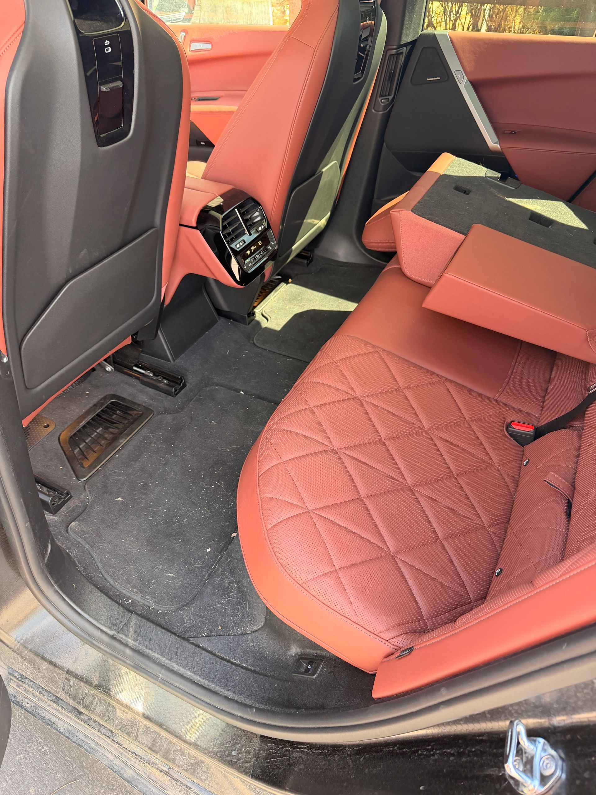 Car interior with red seats and folded seat backs, showing messy floor mats and open door.