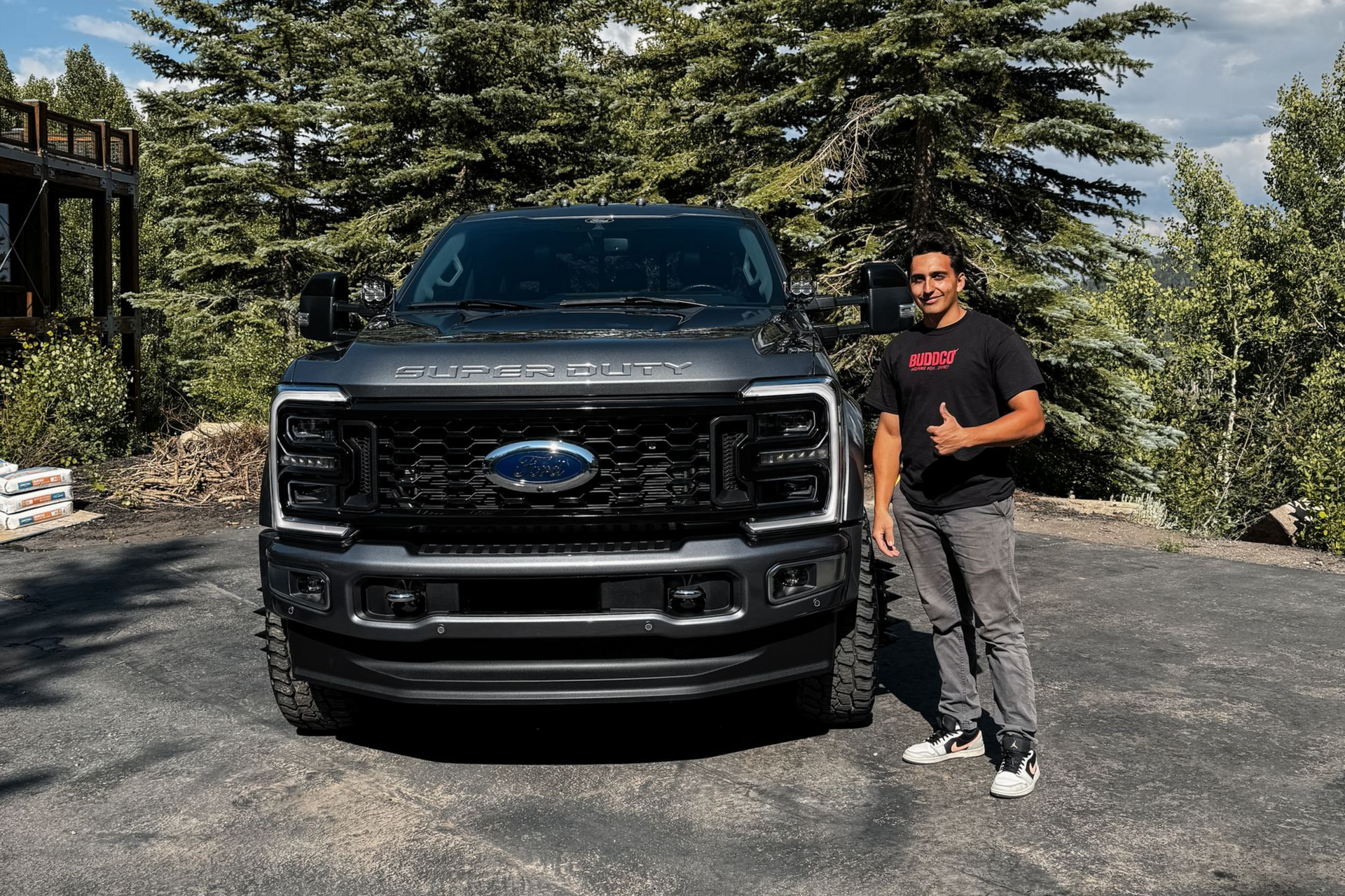 Man standing beside a black Ford truck in a driveway, smiling with trees and a house in the background
