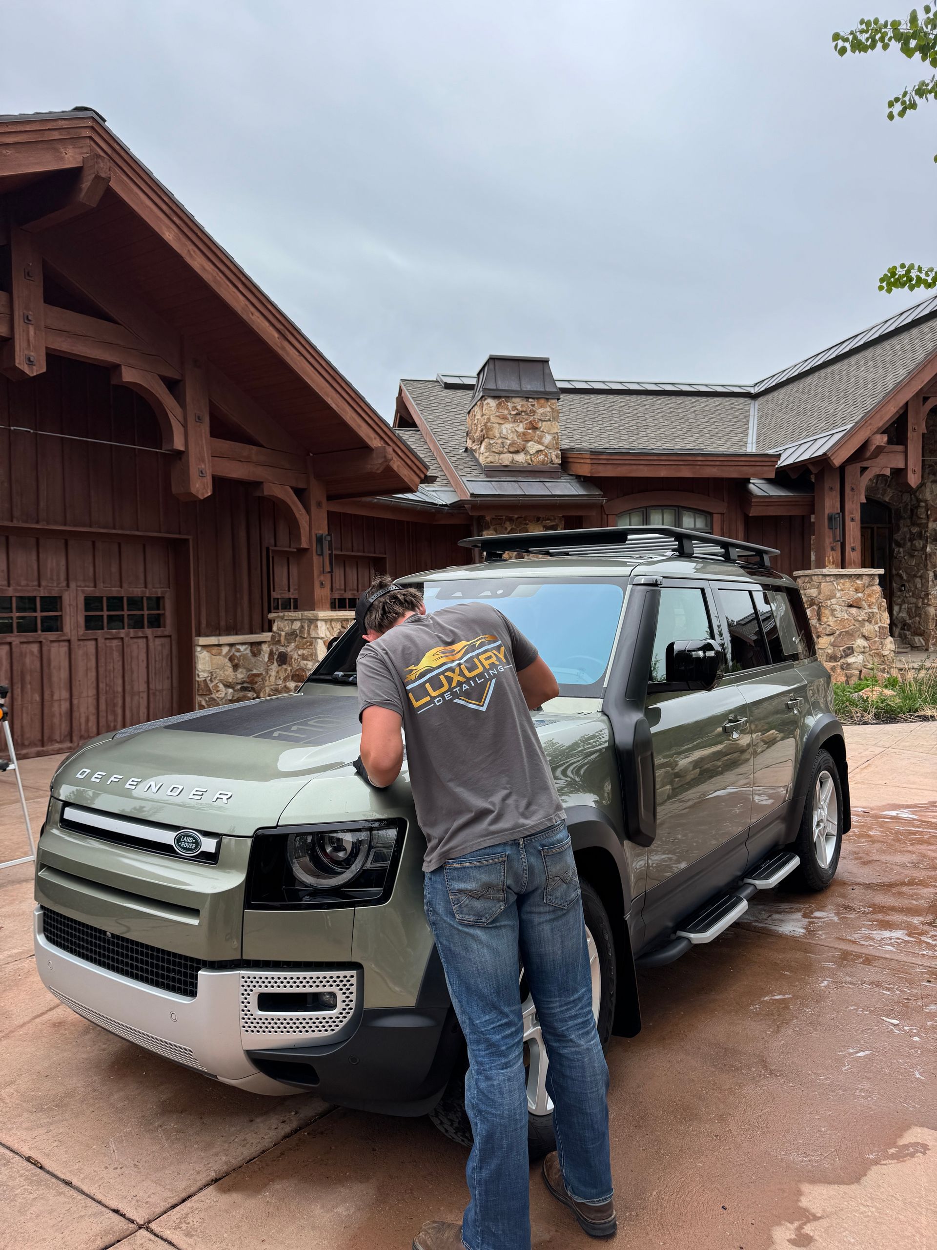 Person cleaning a green SUV in a rustic driveway under an overcast sky.