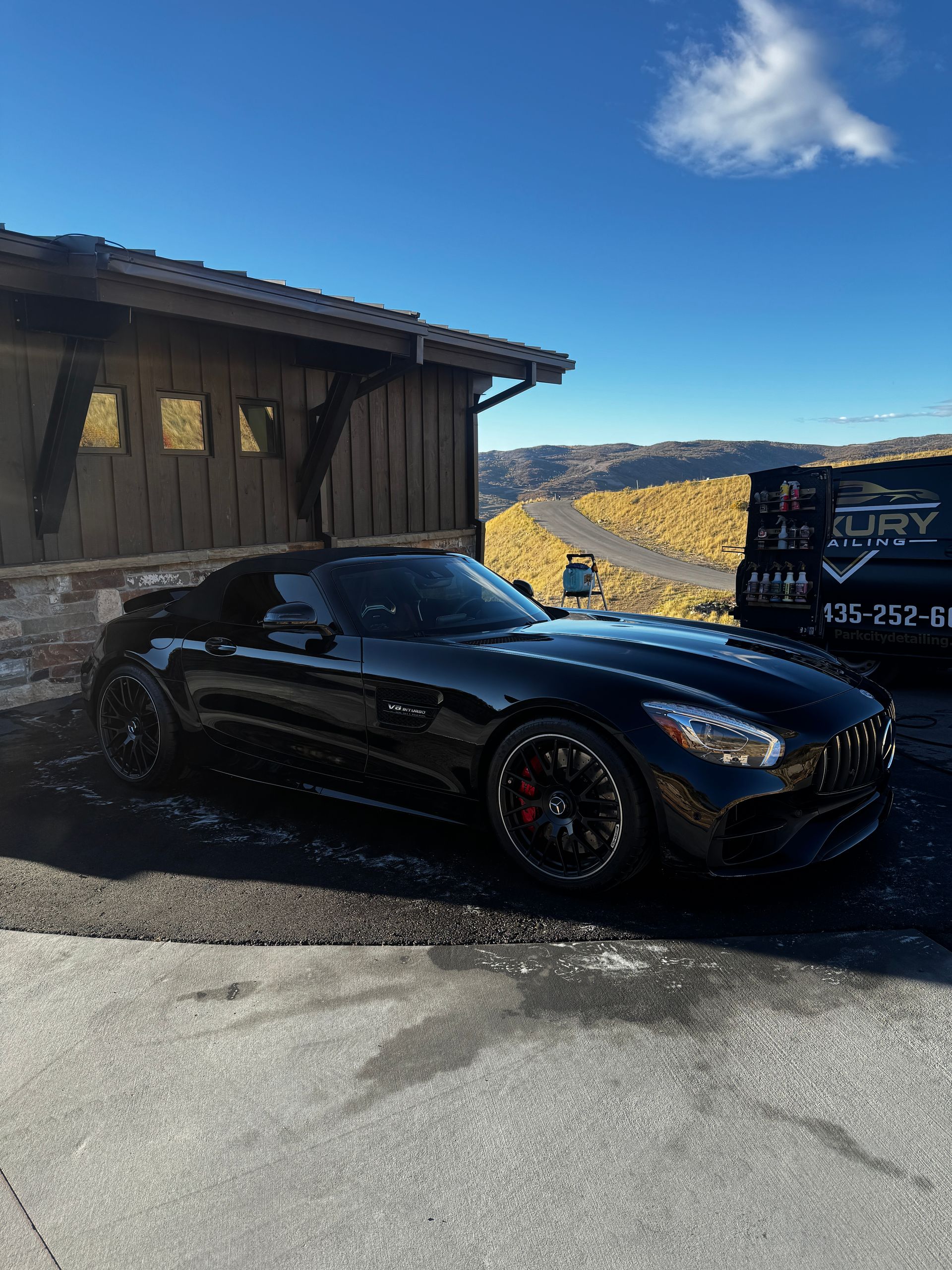 Black sports car parked beside a wooden building on a sunny mountain roadside with snow patches