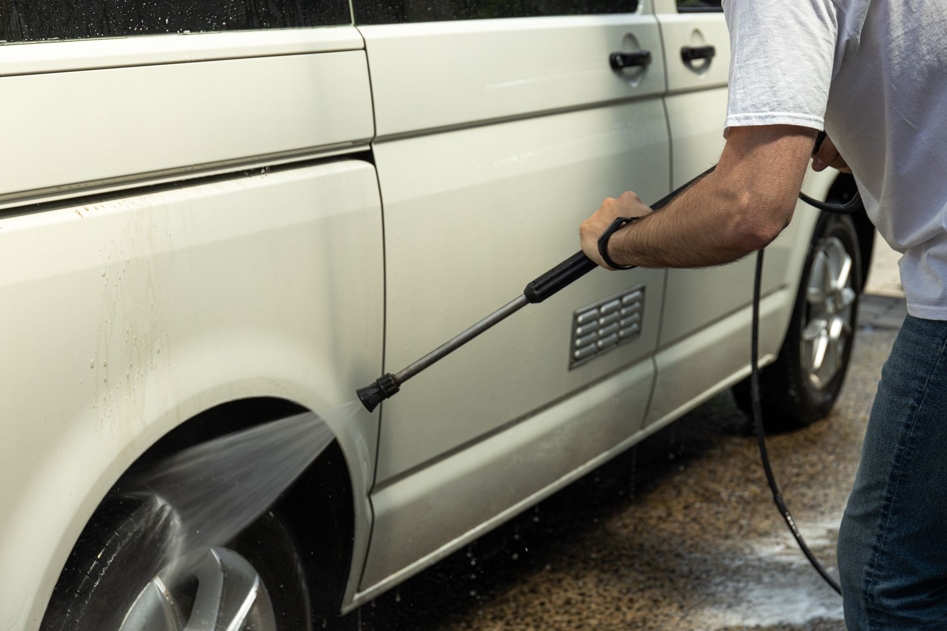 Person pressure-washing a silver van’s side panel and wheel on a driveway