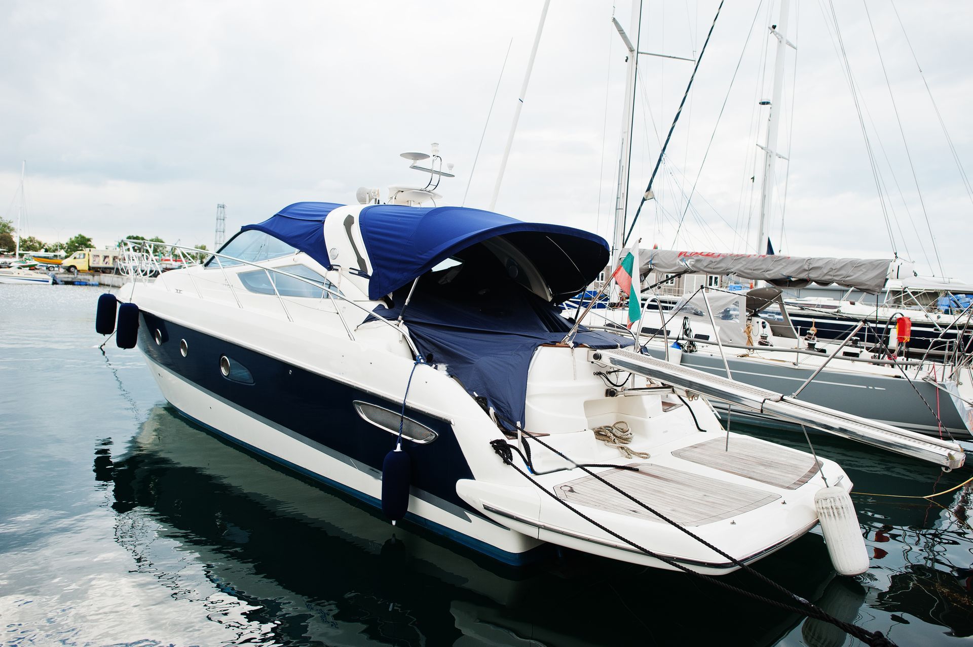 White and blue yacht docked in a marina with calm water and sailboats in the background