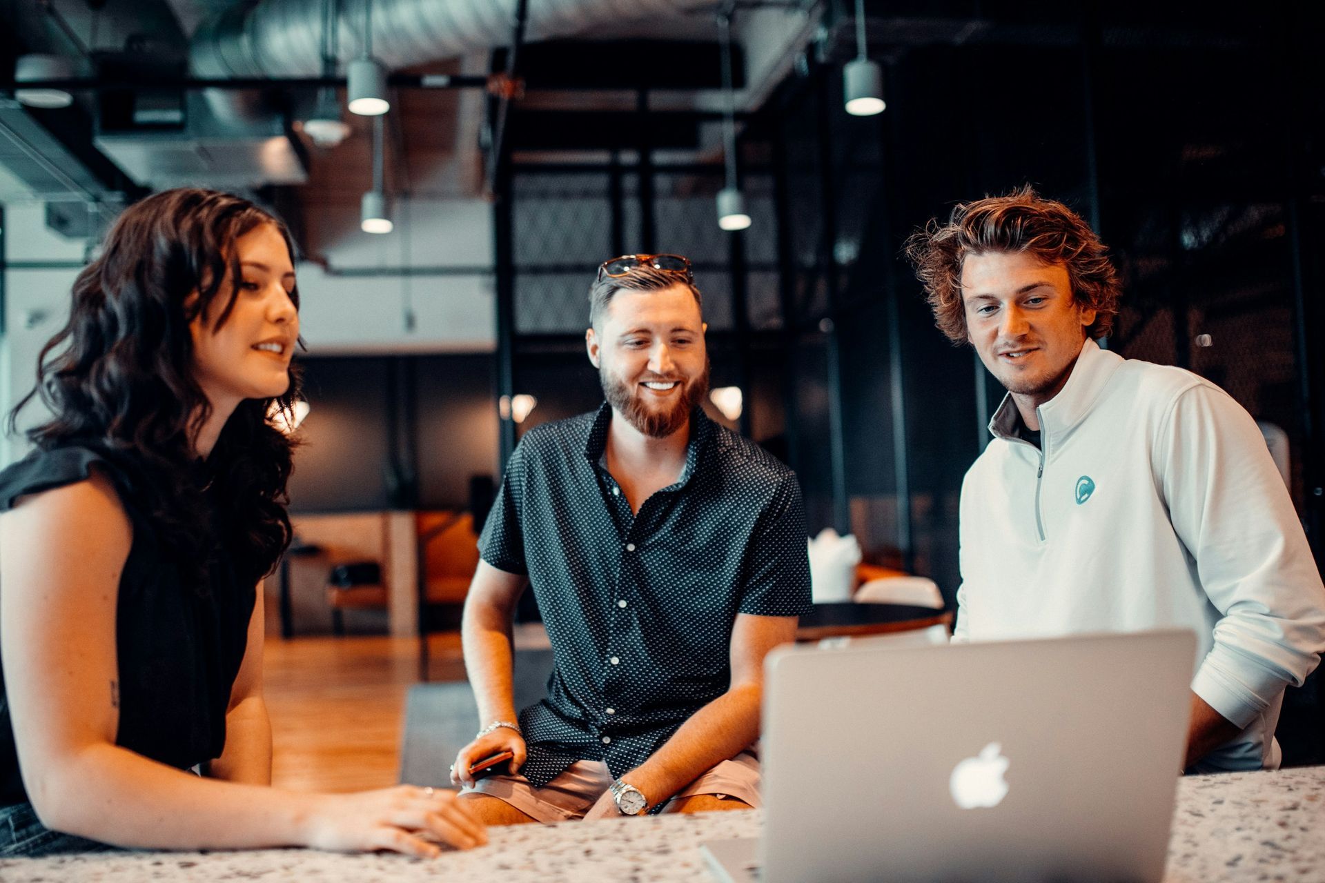 Three coworkers chatting around a laptop in a warm-lit office lounge