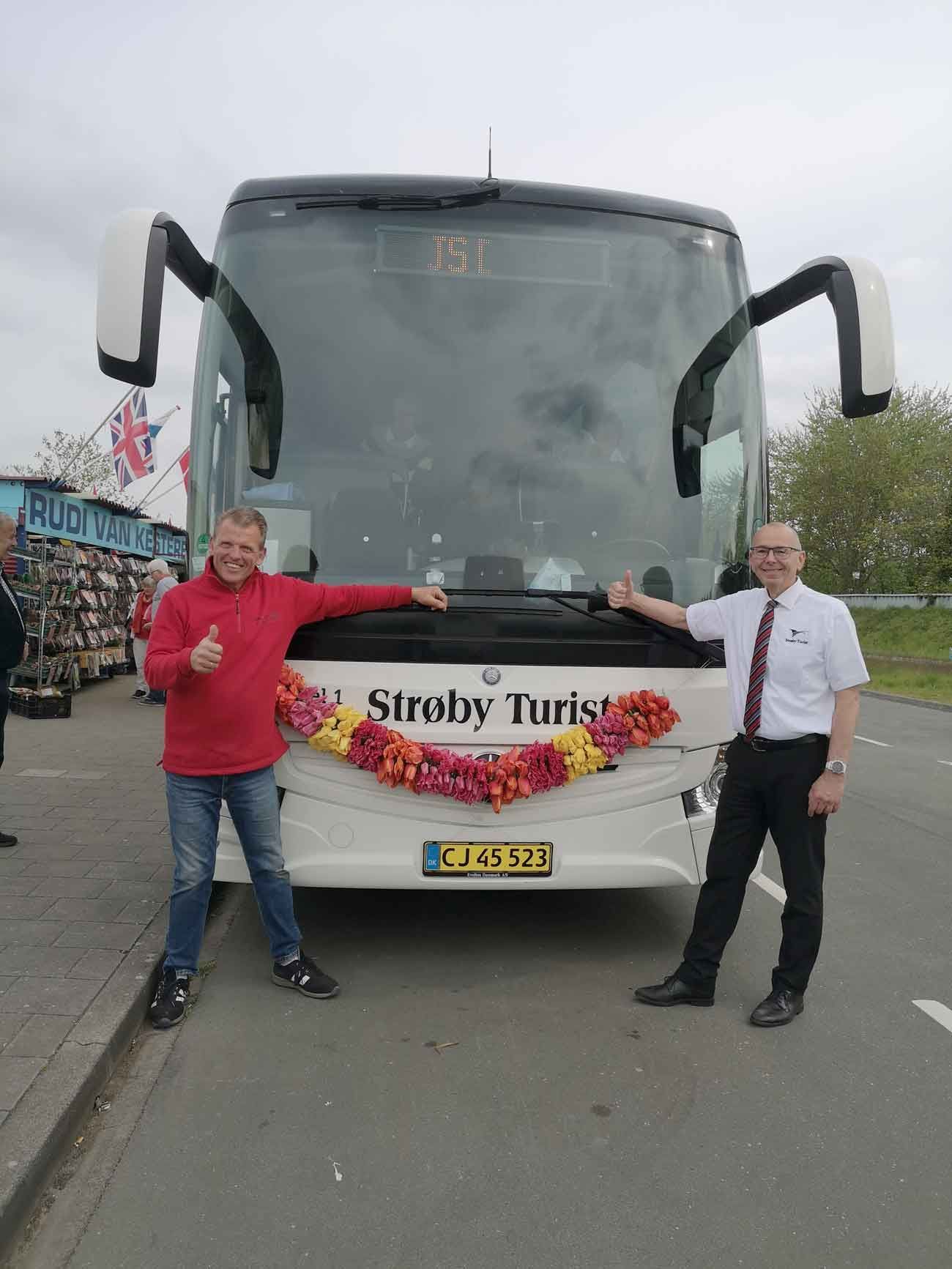 To mænd poserer foran en hvid bus dekoreret med blomster. Mand med røde tommelfingre opad, mand i hvidt smiler.