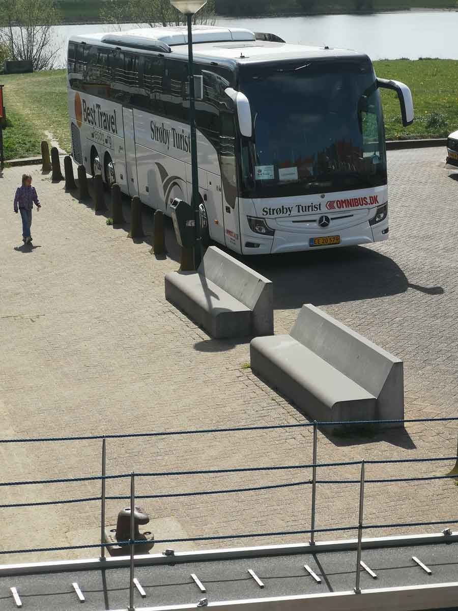 Hvid turistbus parkeret nær betonbænke; en person går forbi.