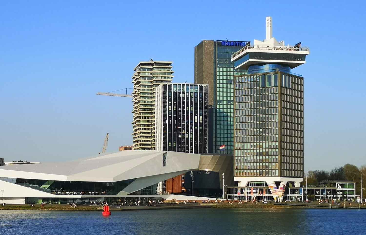 Amsterdams skyline med moderne bygninger, herunder et tårn med et observationsdæk på taget over vandet under en blå himmel.