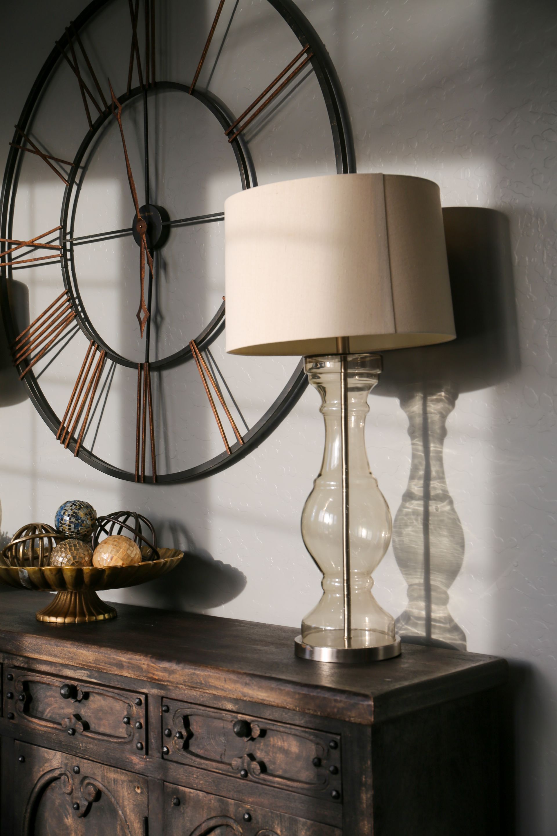 Rustic dresser with two glass lamps and large decorative wall clocks against a gray wall
