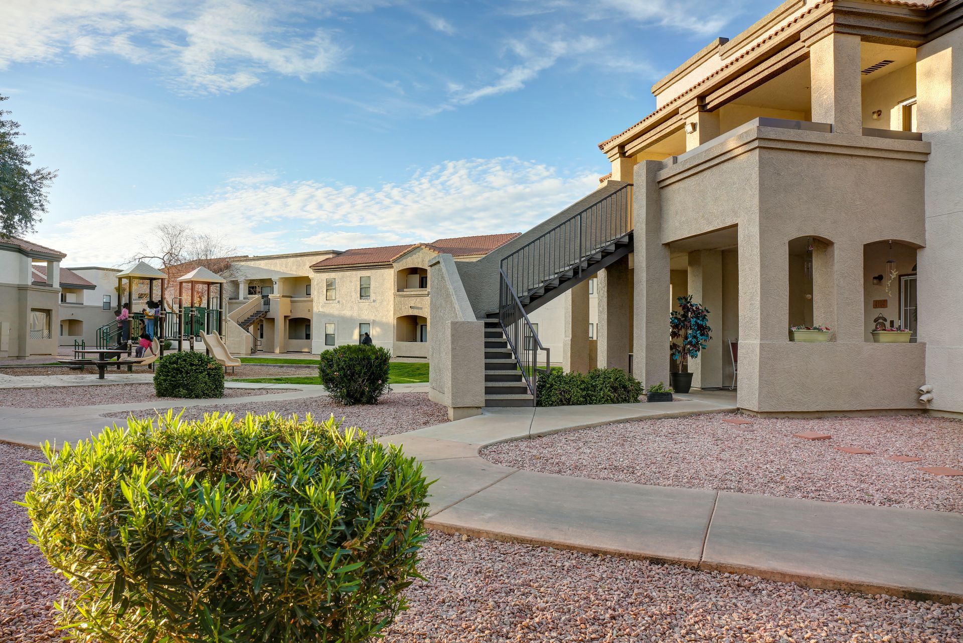 Residential courtyard with beige apartment buildings, exterior stairs, and trimmed shrubs under a blue sky