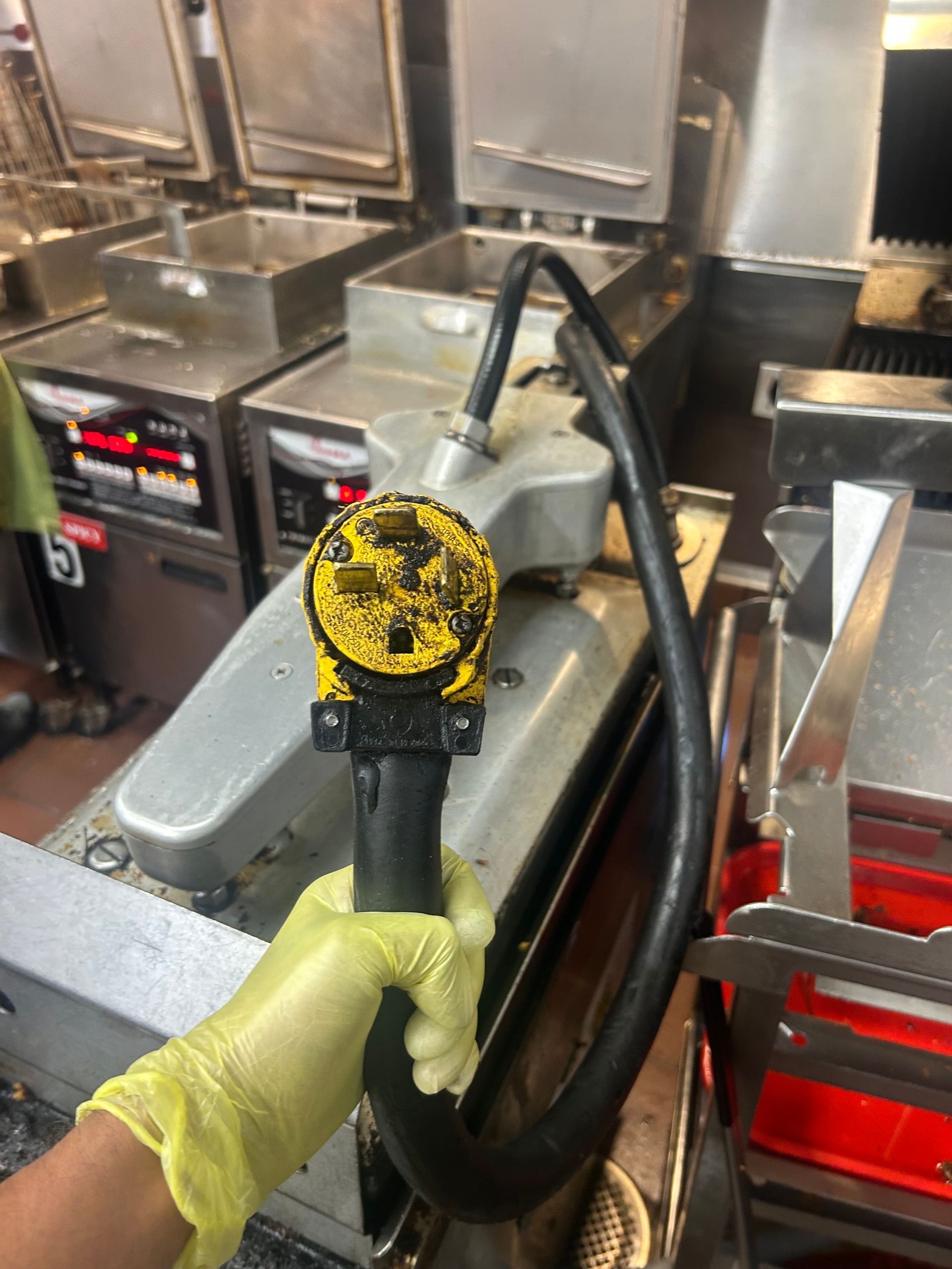 A gloved hand holds a black hose with a yellow connector near kitchen fryers.