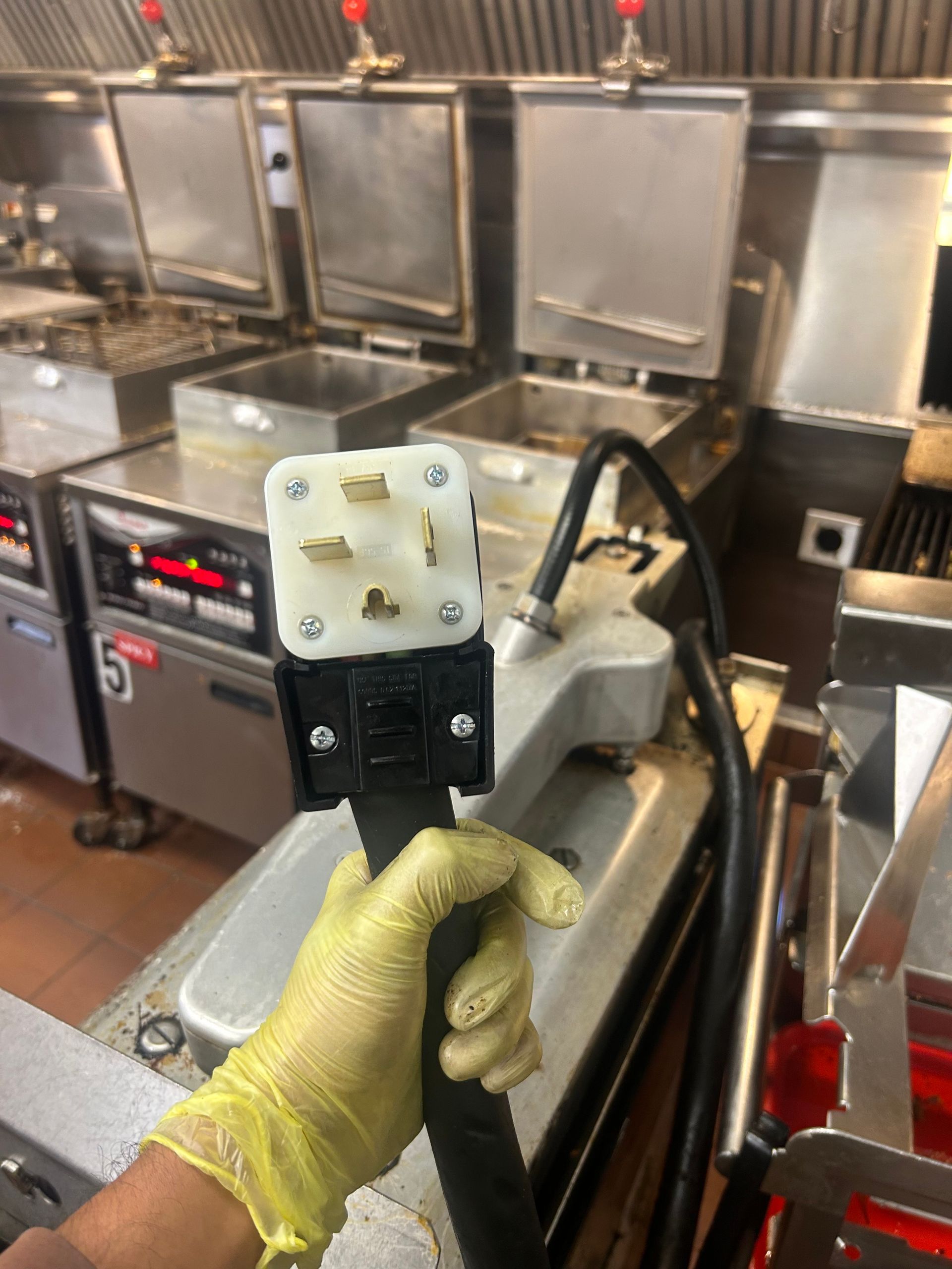 Hand holding a large, four-prong electrical plug in a commercial kitchen with fryers in the background.